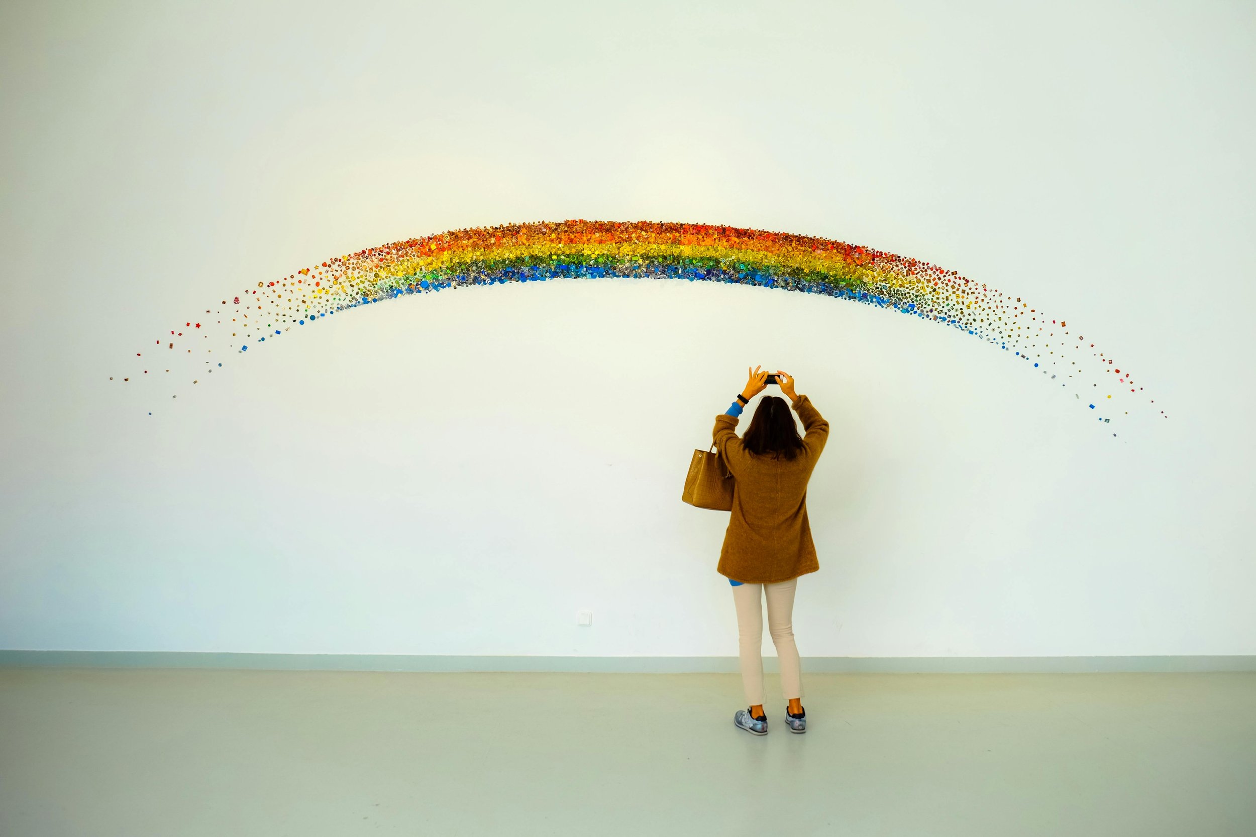 A woman standing in front of a large white wall, taking a photo of a rainbow mural made of small colored tiles arranged in an arc. She is dressed in a brown coat, white pants, and grey sneakers, with a brown bag on her shoulder.