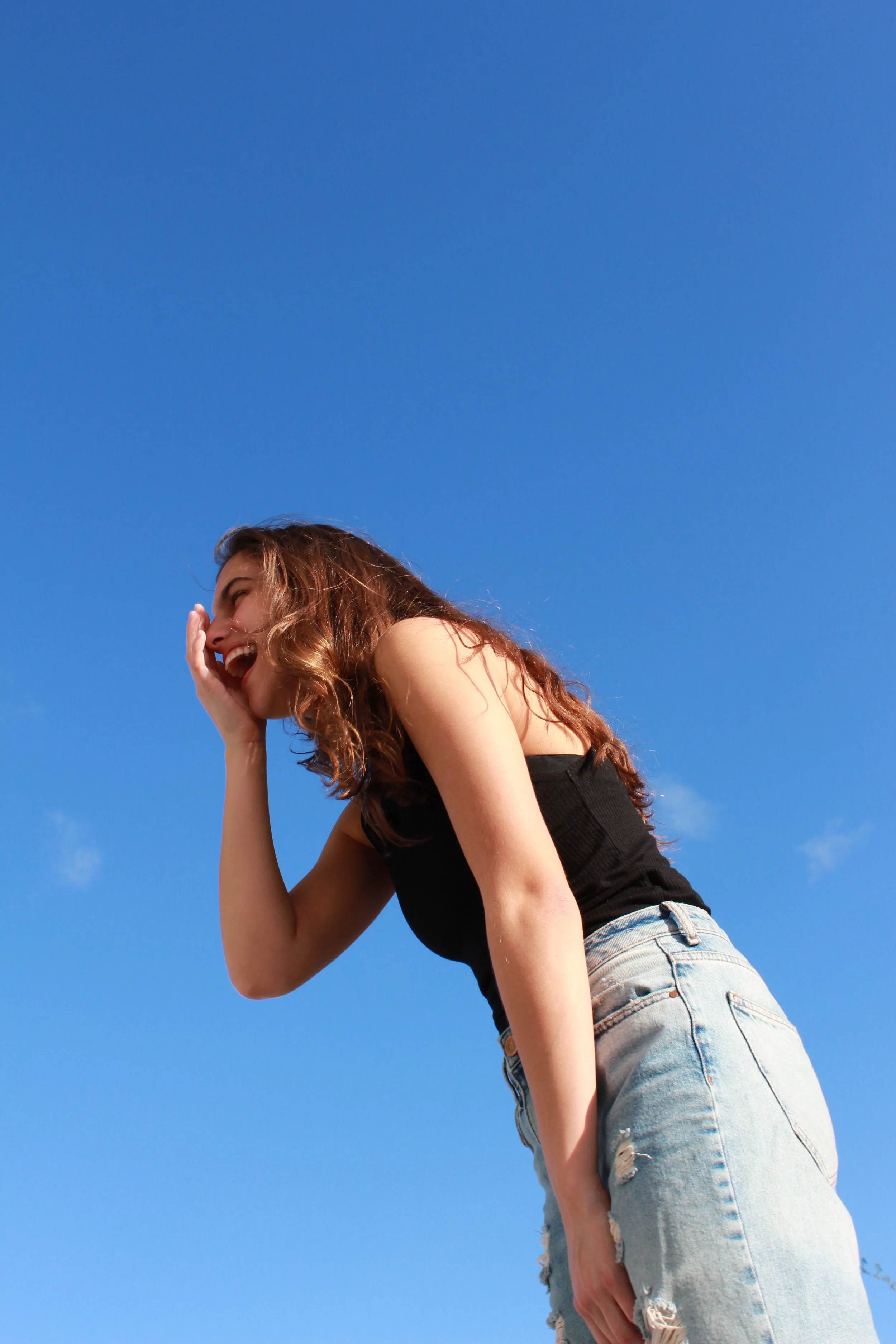 A young woman with curly brown hair laughing outdoors under a clear blue sky, wearing a black sleeveless top and ripped light blue jeans.