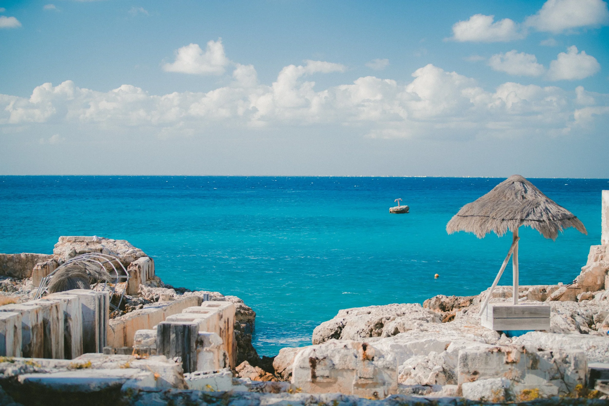 A sunny beach scene with a thatched umbrella on rocks, turquoise ocean, and a boat in the distance under a partly cloudy sky.