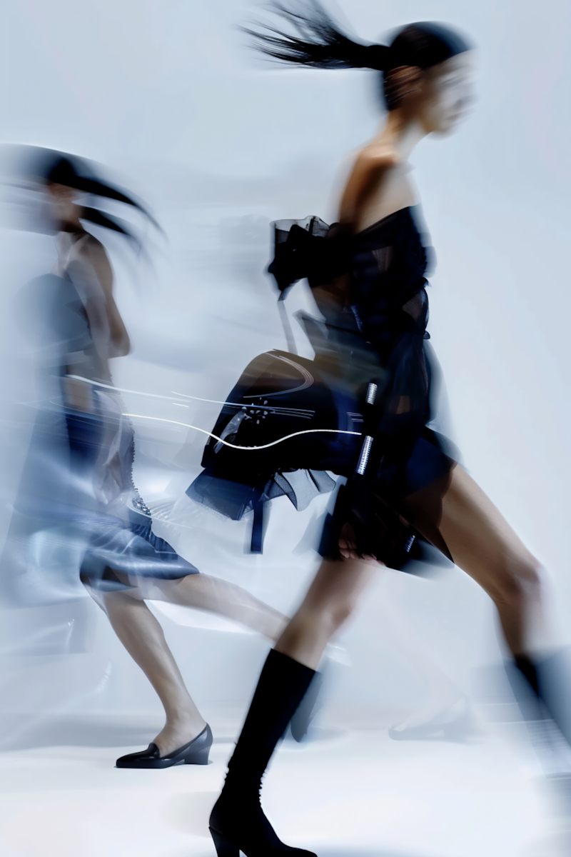 Two women walking quickly, one in black boots and the other in heels, with motion blur and wind blowing their hair.