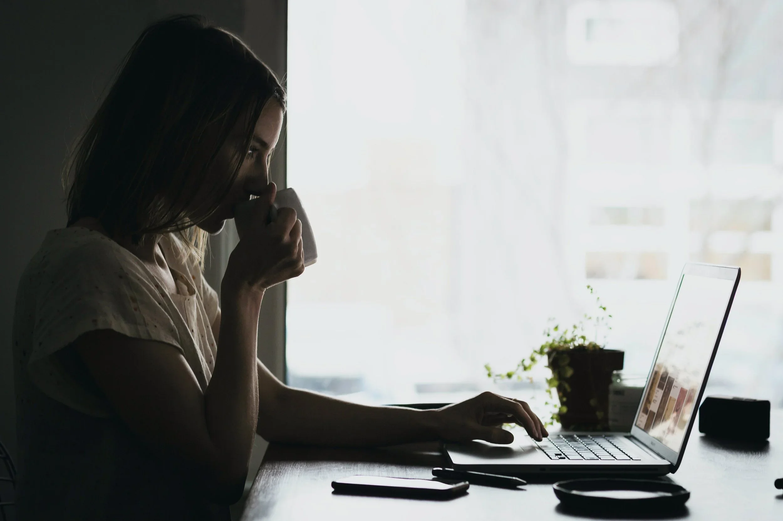 A woman with shoulder-length hair drinking from a mug while working on her laptop at a desk, near a bright window with sheer curtains, with a small potted plant and phone on the desk.