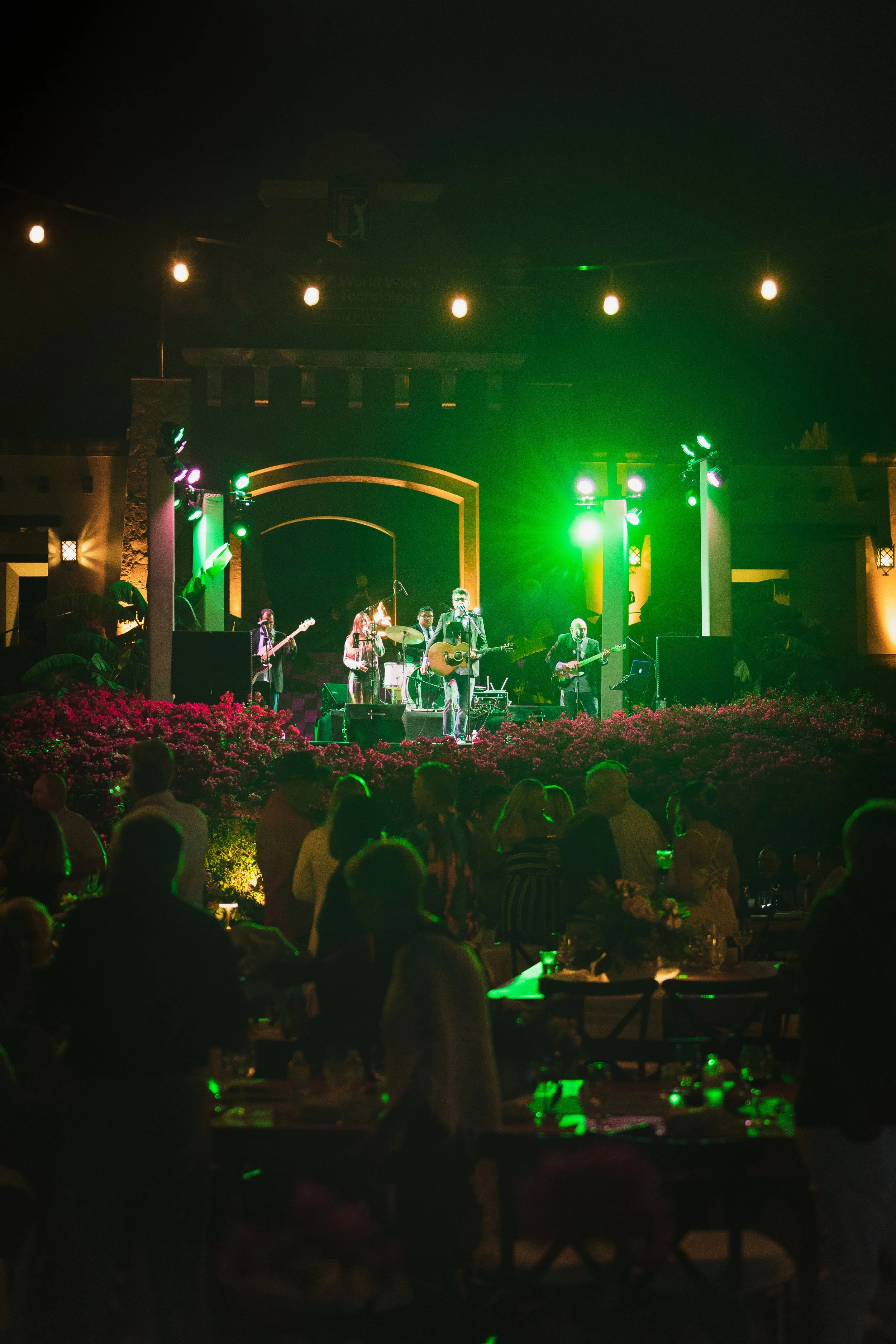 Nighttime outdoor concert with a band on stage under green lighting, audience members standing and sitting at tables, and string lights overhead.