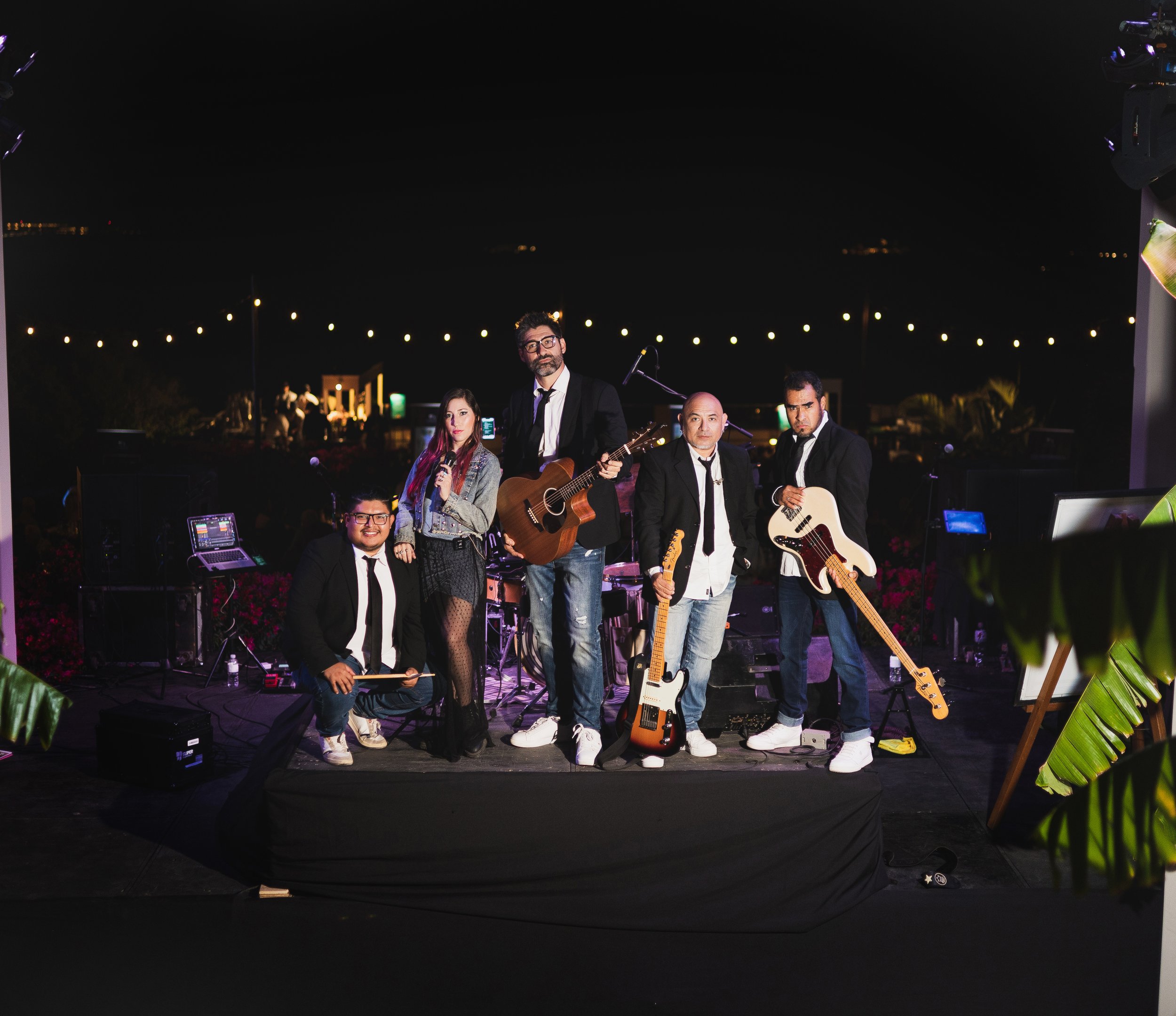 A group of five musicians performing on stage outdoors at night, with instruments including guitars and a microphone, under string lights, surrounded by foliage.