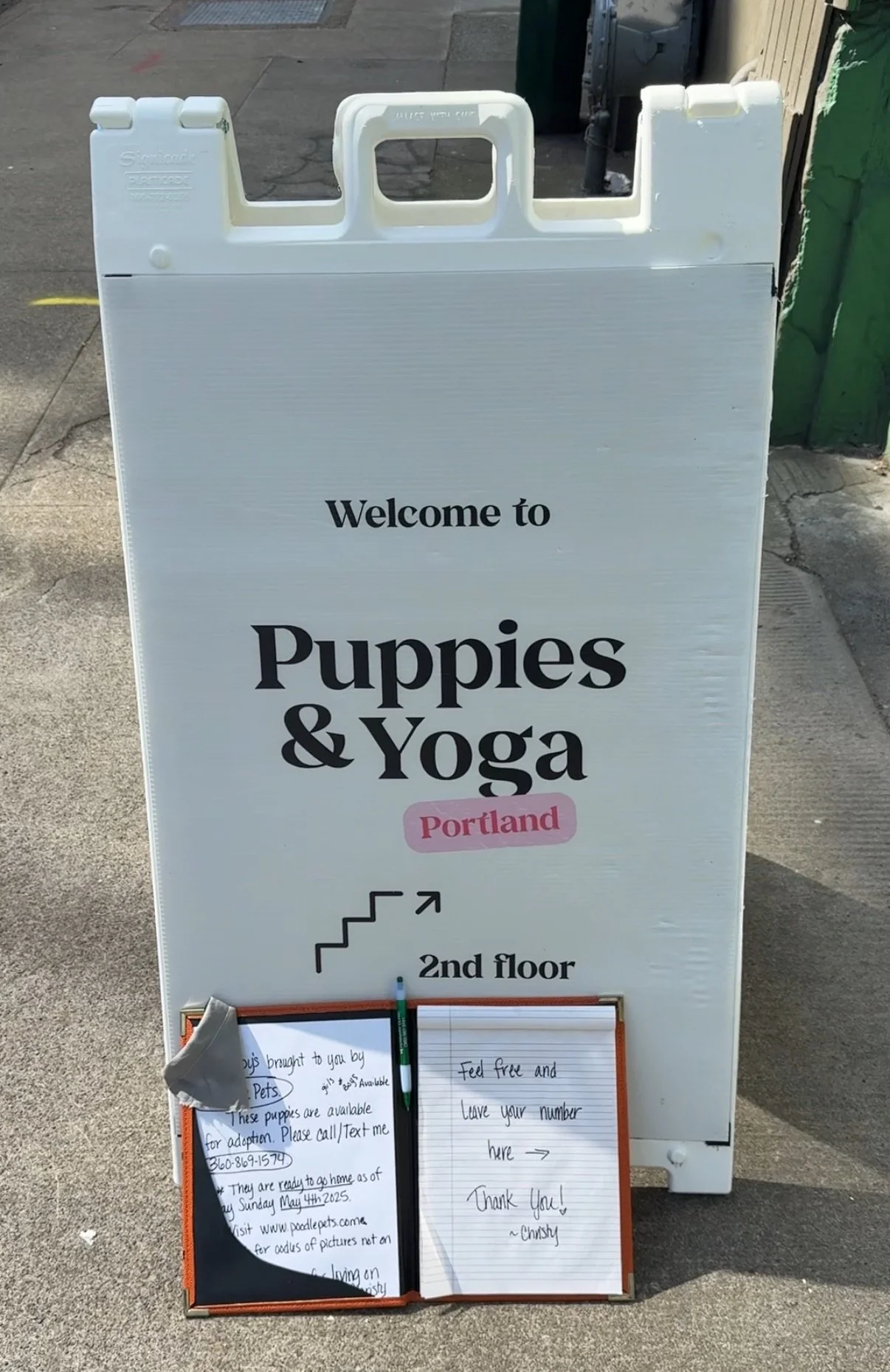 A white sandwich board sign on the sidewalk advertising Puppies & Yoga in Portland on the 2nd floor, with handwritten notes and a marker inside two clipboards at the bottom.