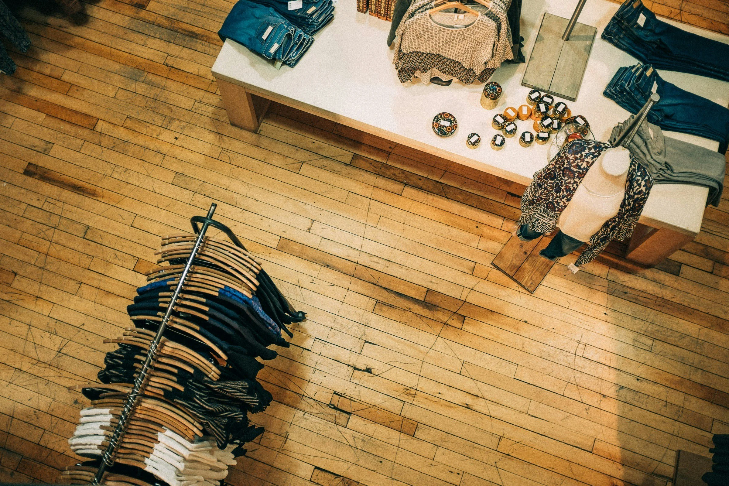 Overhead view of a retail clothing store display with folded jeans on a white table, neatly arranged stacks on racks including shirts, sweaters, and pants, on a wooden floor.