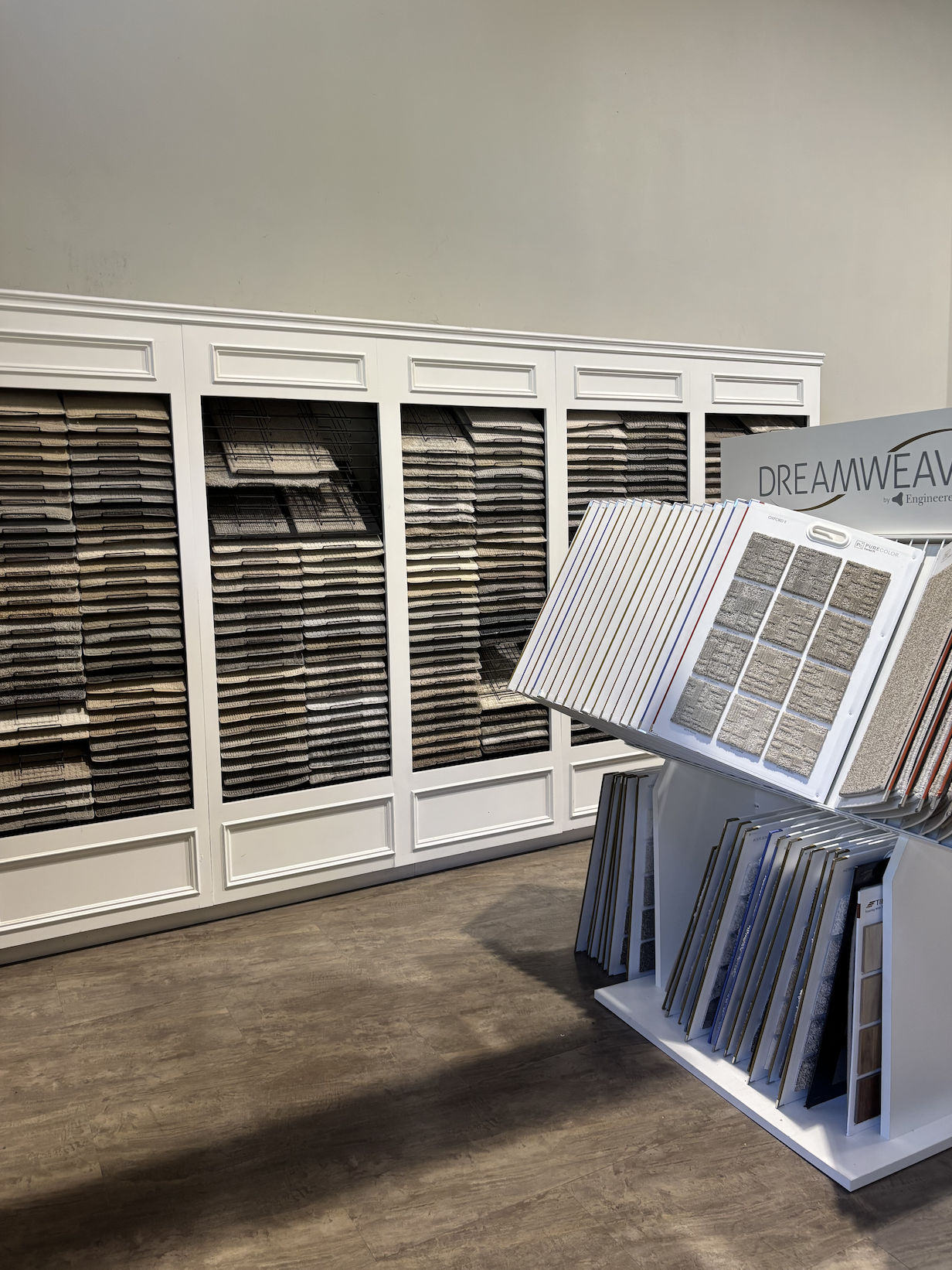 Flooring samples displayed in a showroom with a white cabinet filled with tile or laminate samples, and an open sample book on a stand showing various flooring options.