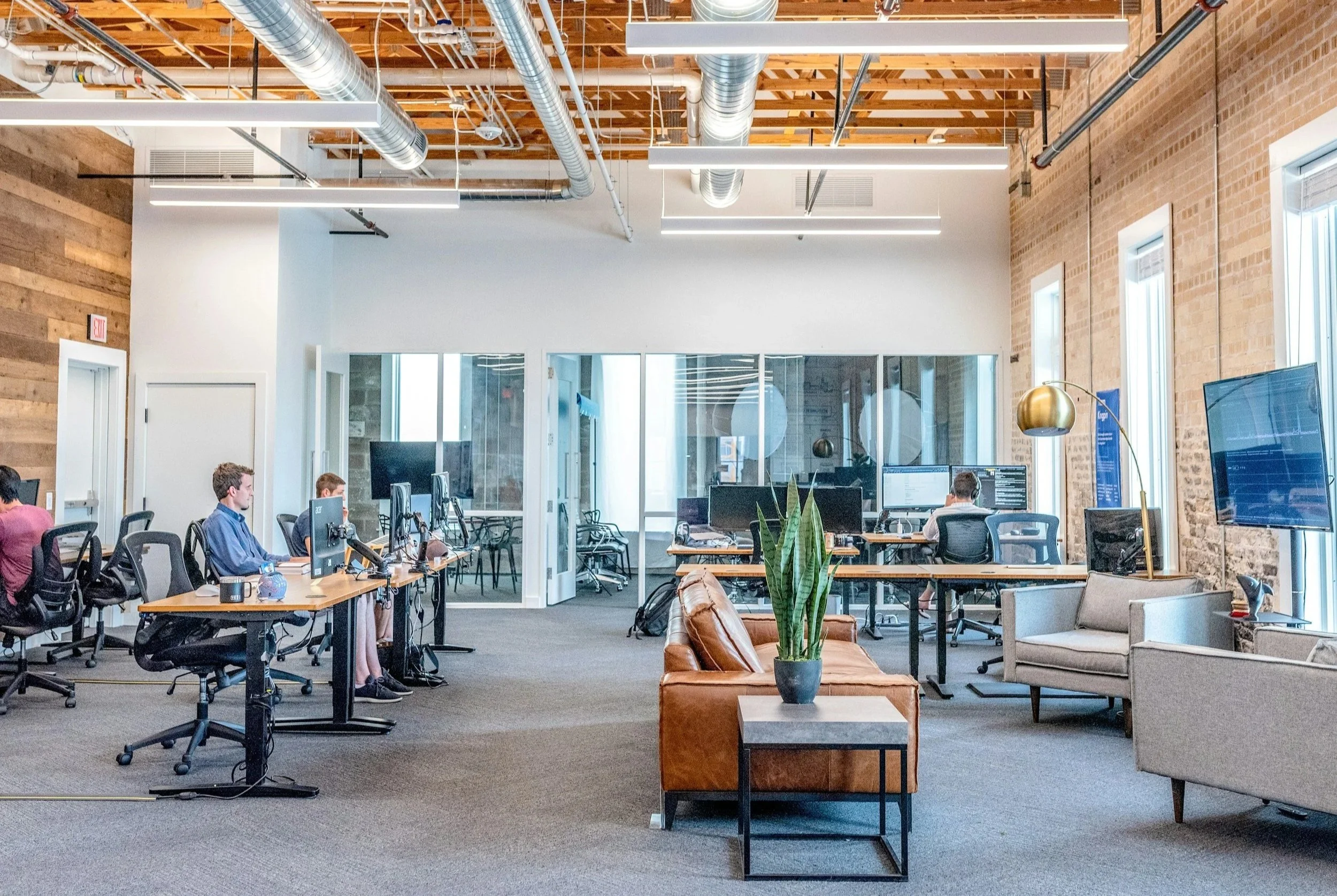 Open-concept modern office with exposed brick and ductwork ceilings, multiple workstations with computer monitors, and a sitting area with leather and fabric chairs and a coffee table with a potted plant.