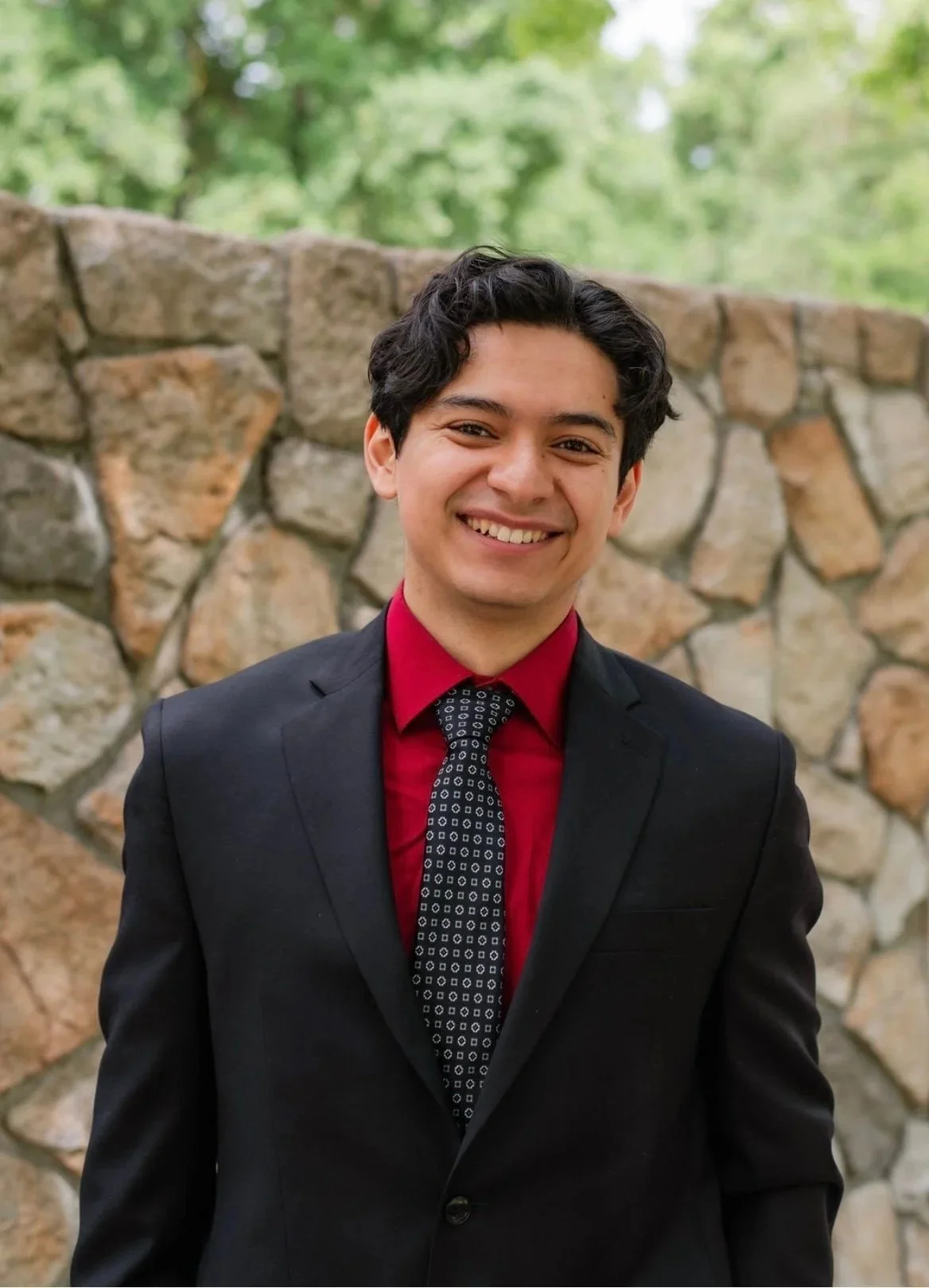 A young man in a black suit, red dress shirt, and patterned tie standing outdoors in front of a stone wall with green trees in the background, smiling.