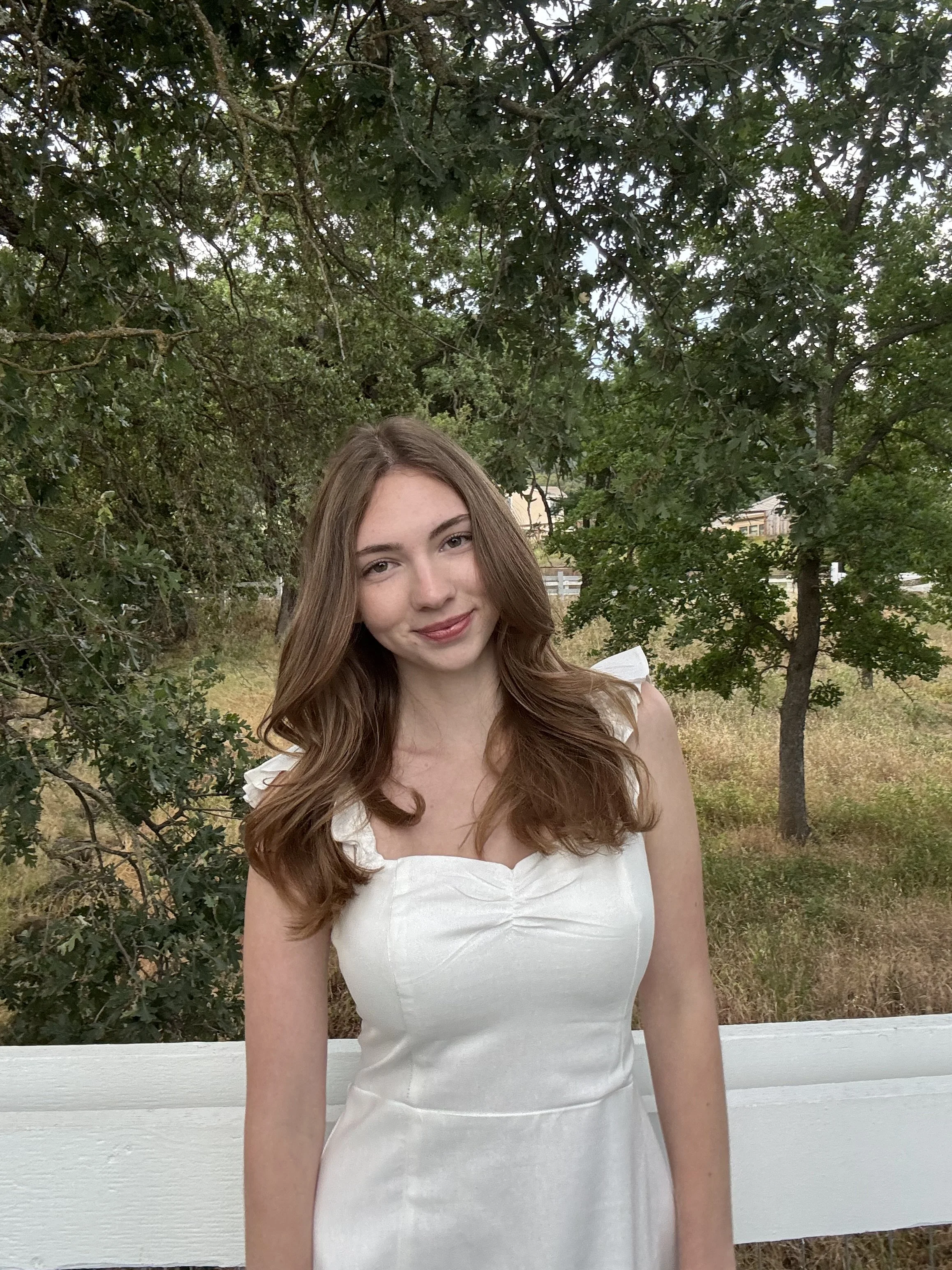 Young woman with long, wavy brown hair smiling in front of trees with green leaves, wearing a white dress.