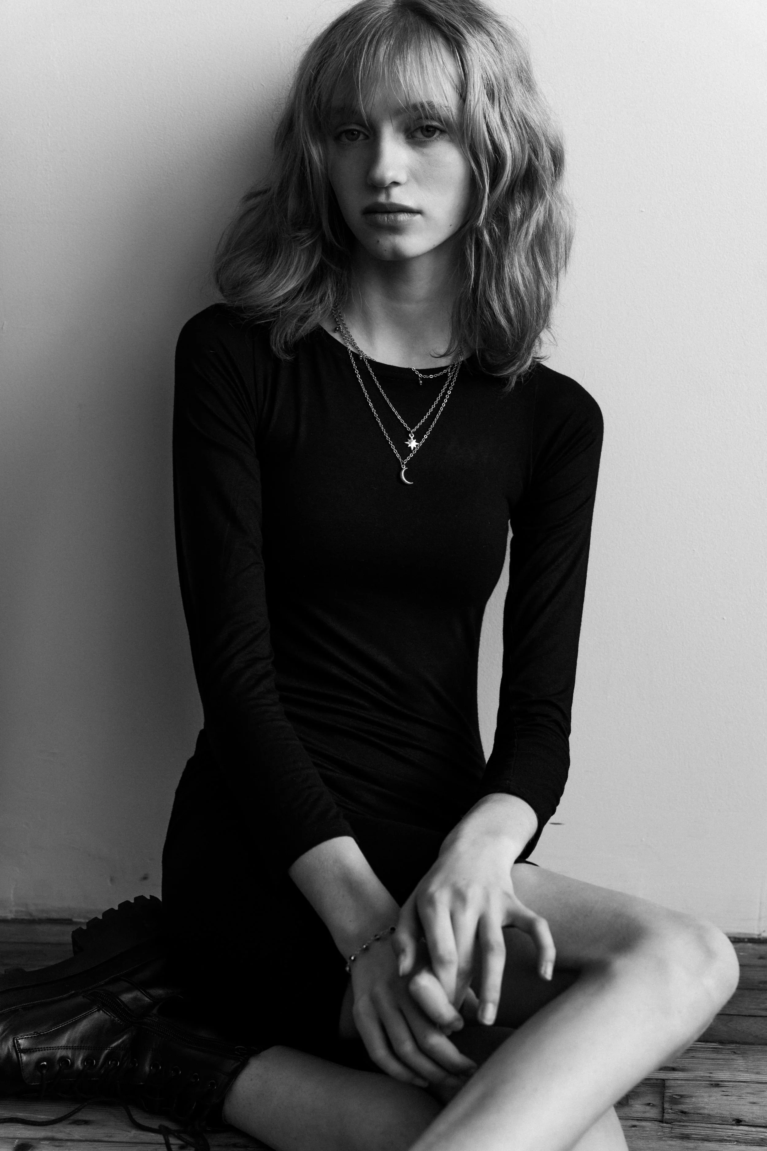 Black and white portrait of a woman sitting with crossed legs, wearing a black dress and layered necklaces; she has wavy hair and a serious expression.