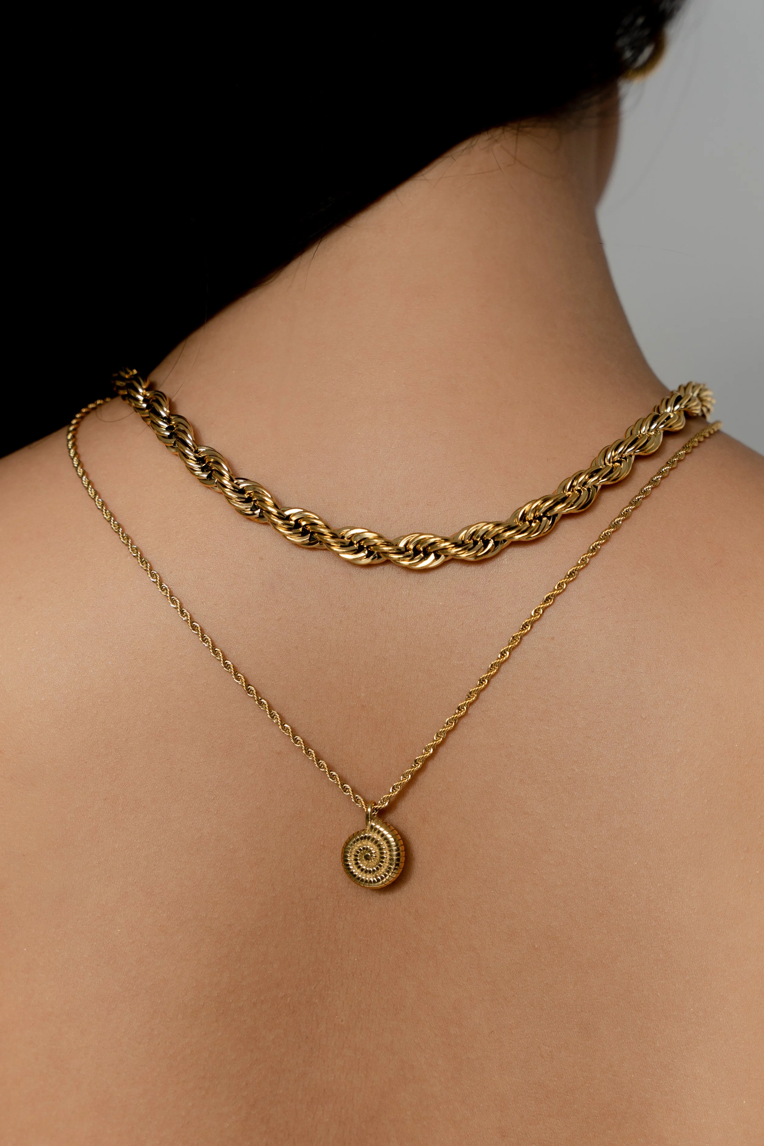 Back view of a person wearing two gold necklaces, one with a twisted chain and one with a circular pendant, against bare skin.