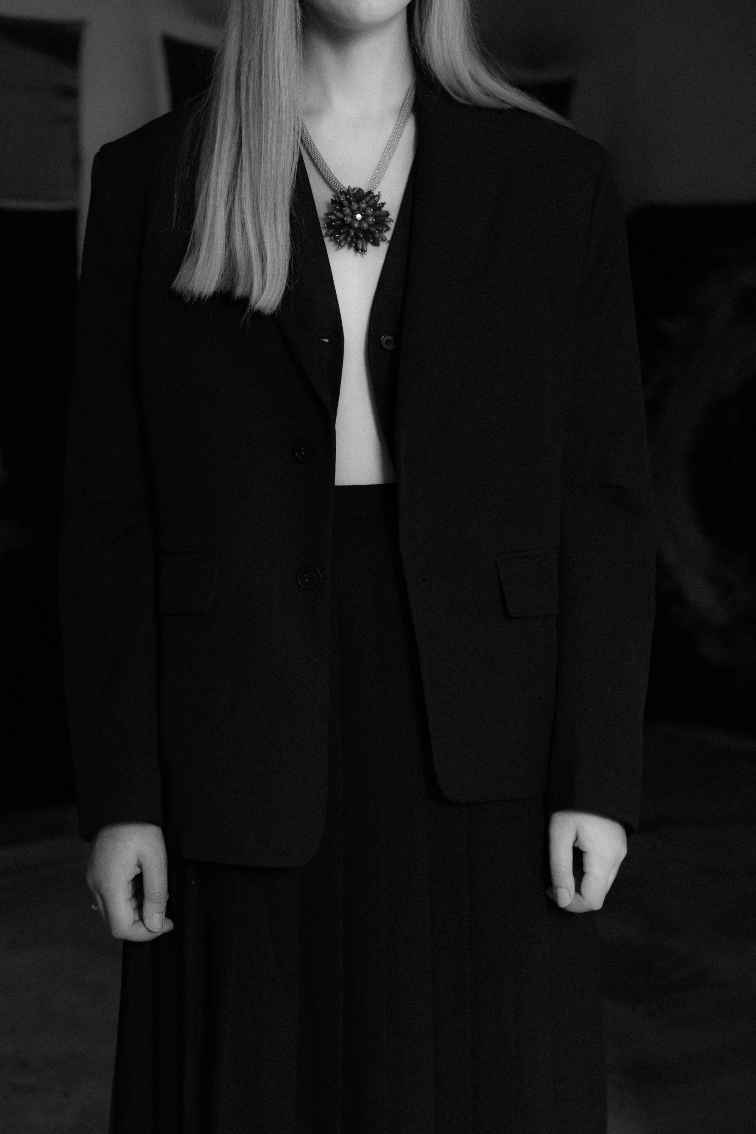 Black and white photo of a person wearing a formal black jacket and a large statement necklace with a floral design.