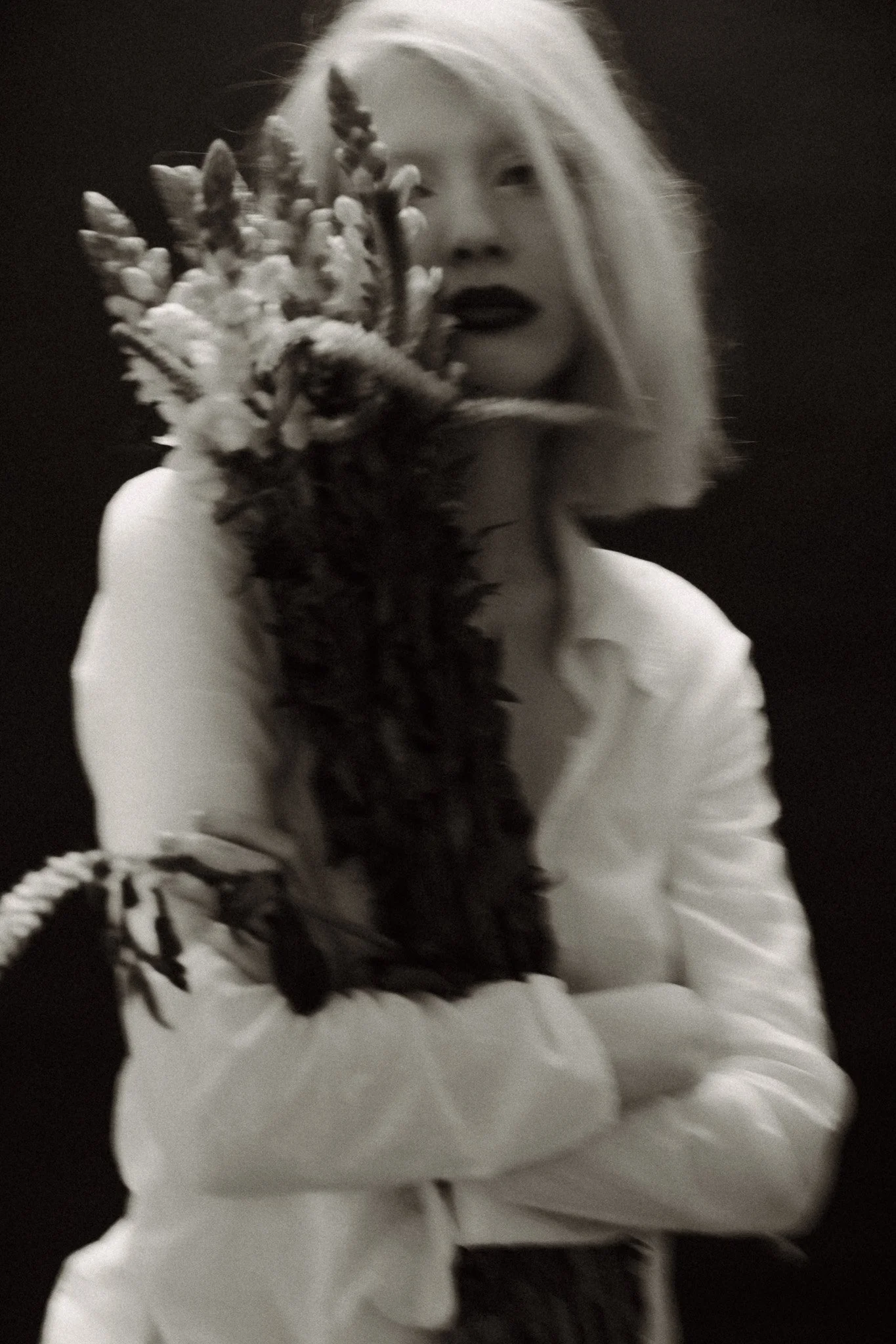 Black and white photo of a person with short blonde hair holding a bouquet of flowers, wearing an open-collared shirt.