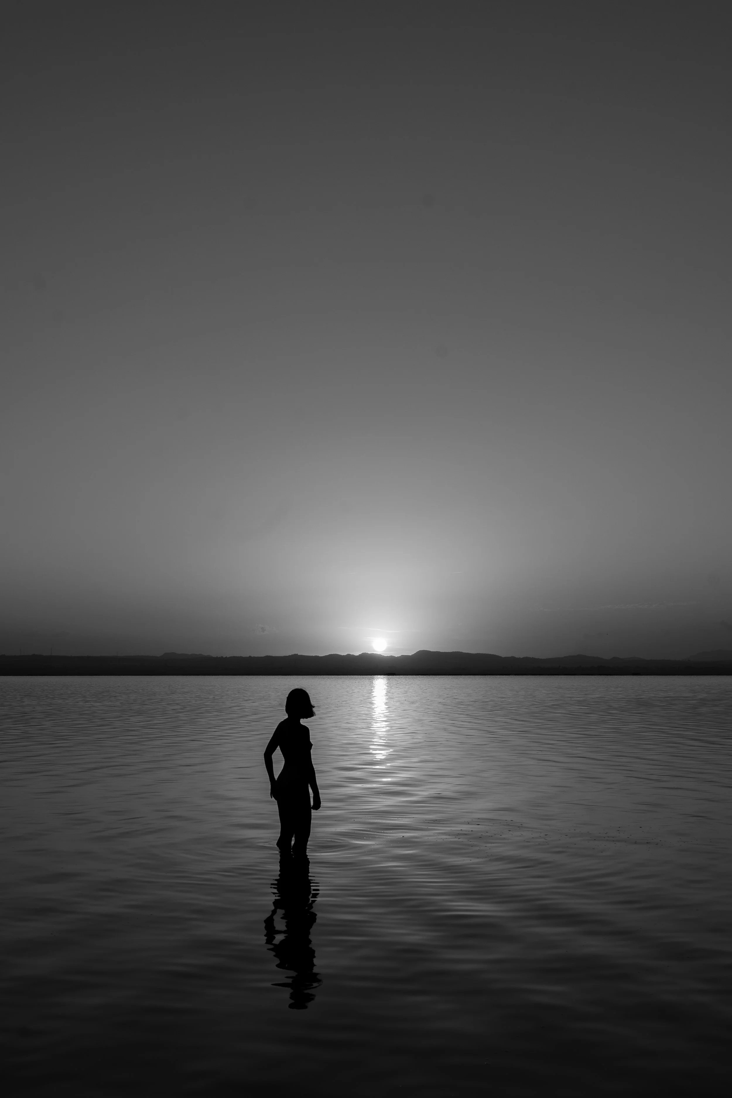 Silhouette of a person standing in water at sunset, with reflections on the surface in a black and white photograph.