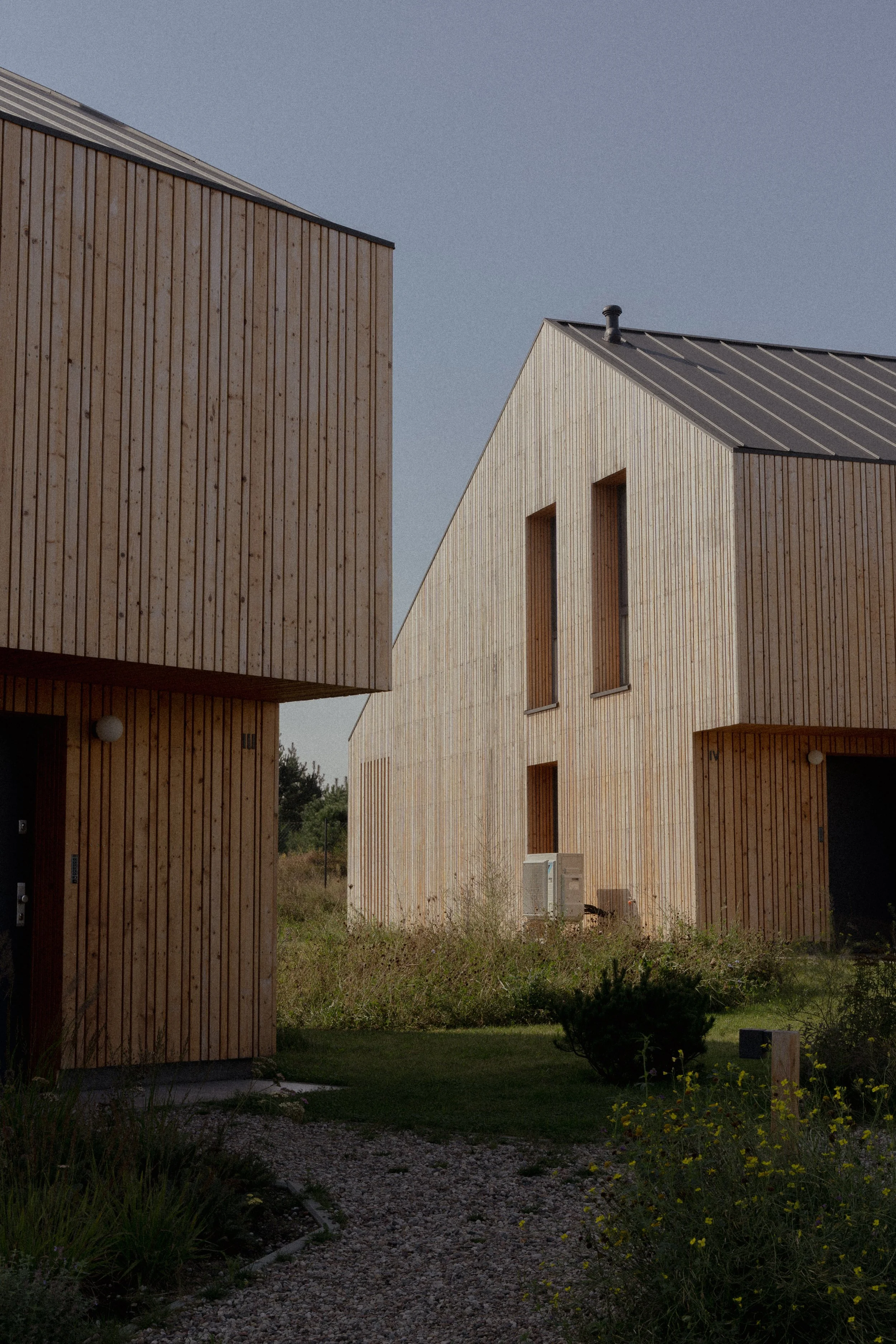 Modern wooden houses with vertical siding and metal roofs, surrounded by grass and plants.