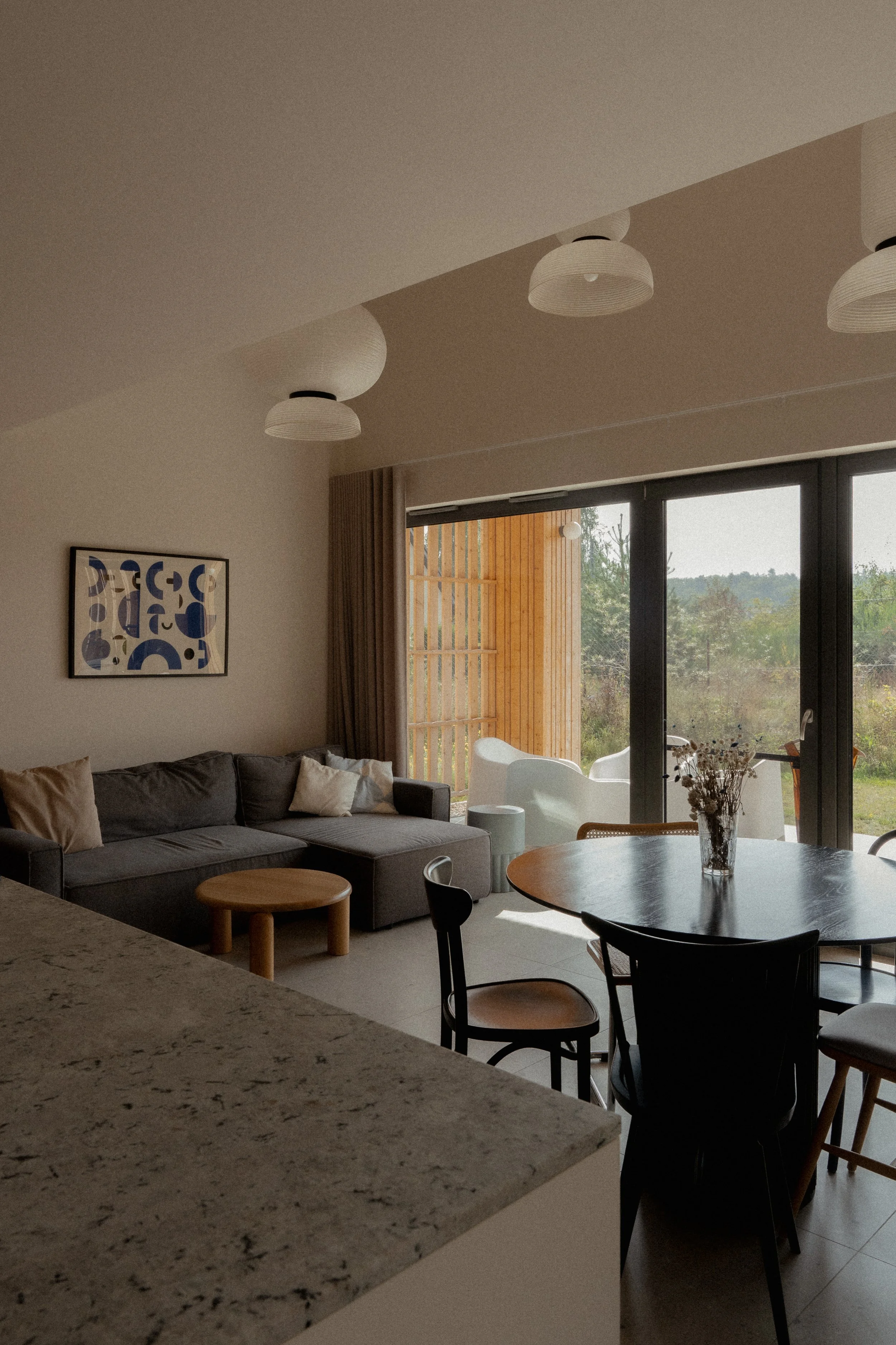 Modern living room with gray sectional sofa, round wooden coffee table, dining table with chairs, abstract wall art, pendant lighting, and large sliding glass doors opening to a deck.