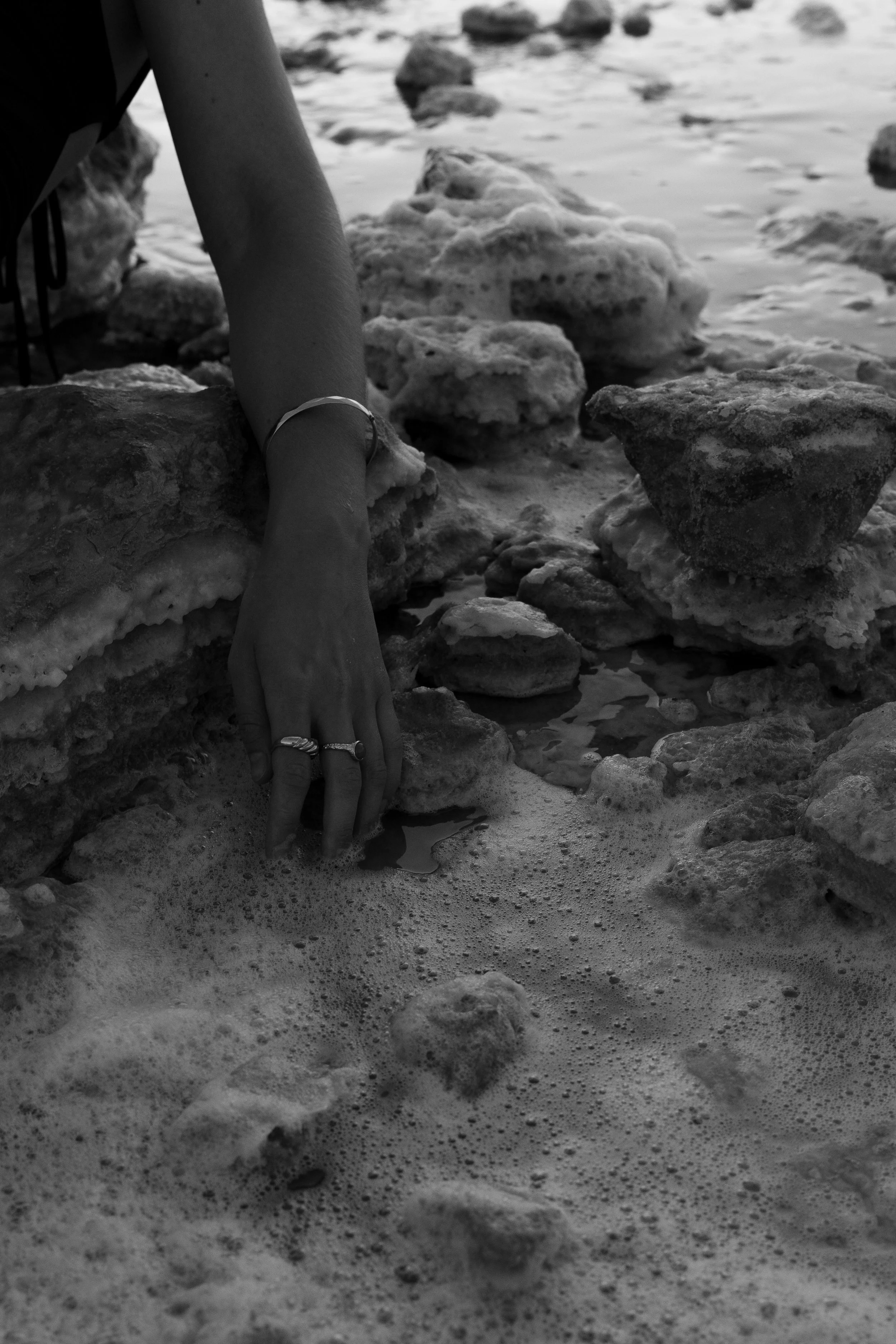 Person's hand touching water among salt-covered rocks and foam.