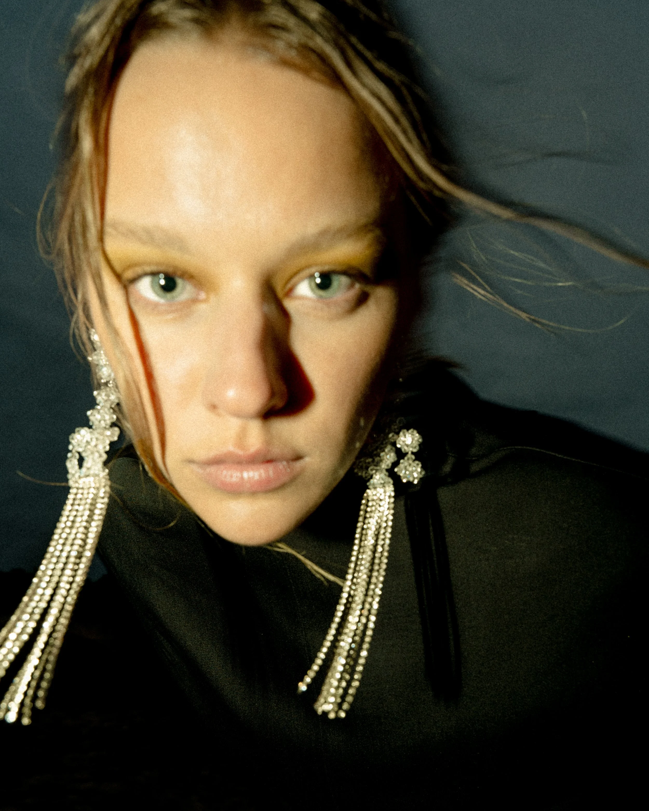 Close-up of a person with long, ornate earrings and an intense expression, wearing a dark top against a dark background.