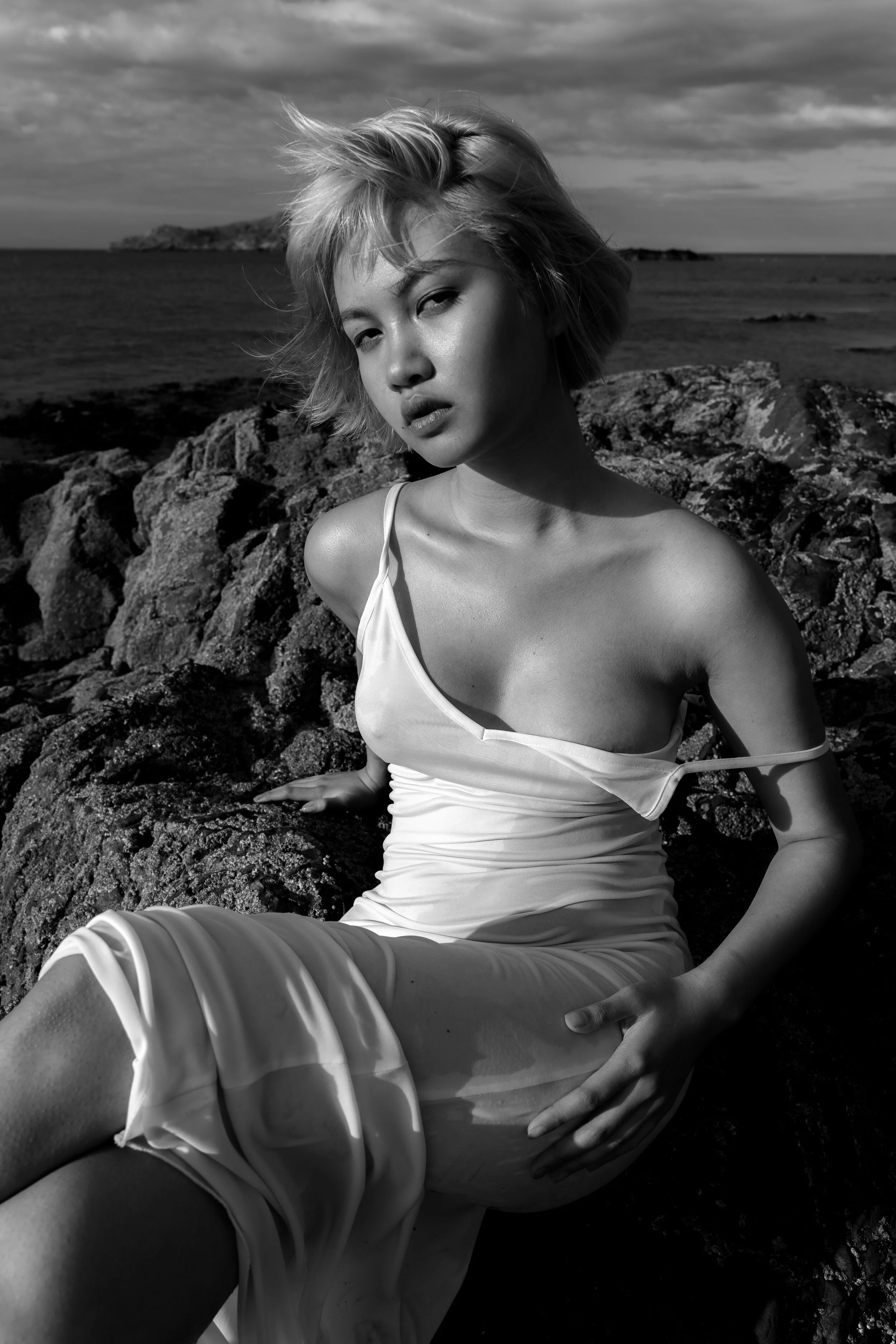 Black and white portrait of a woman in a white dress sitting on rocks by the sea.