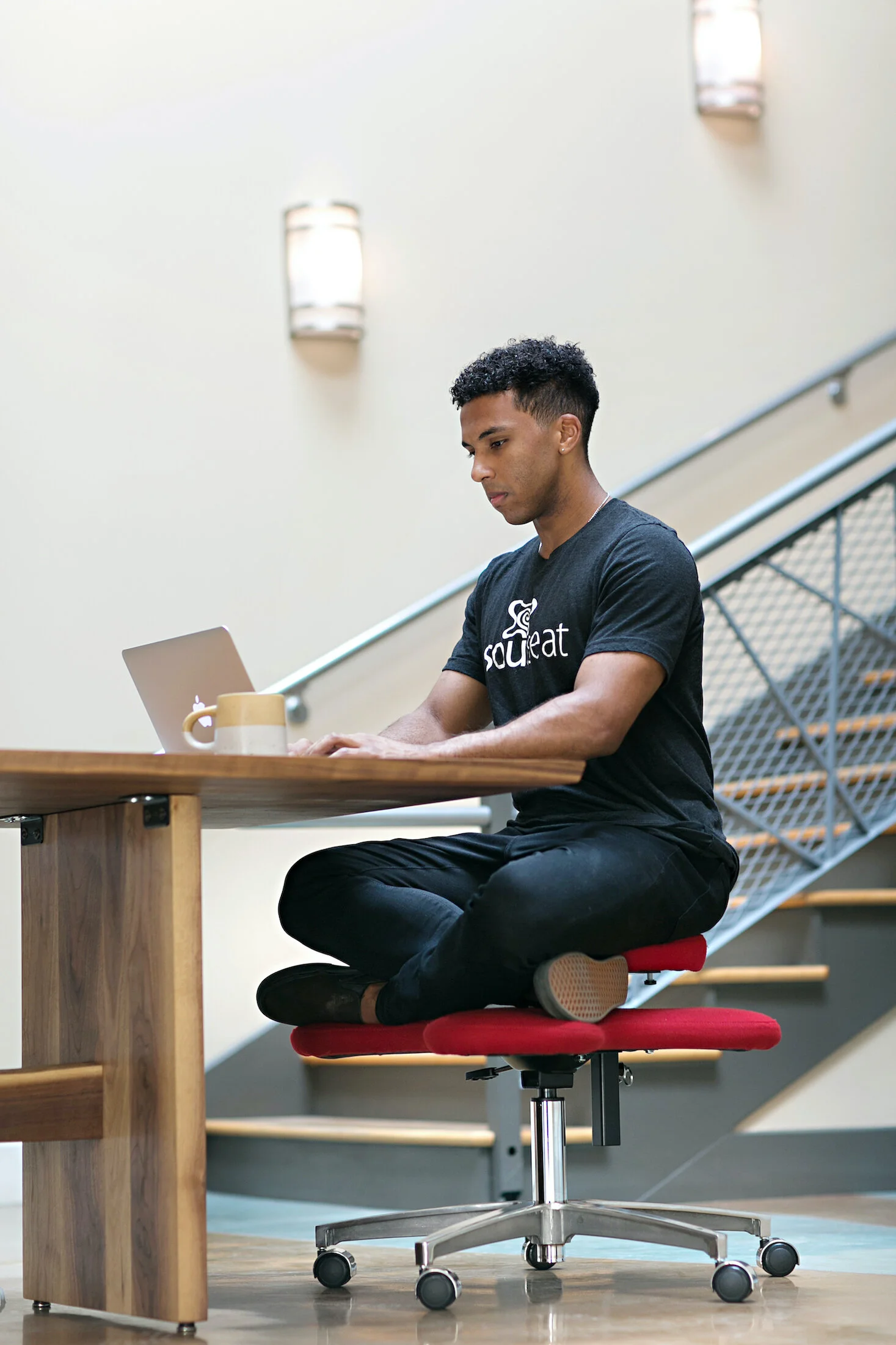 A young man with dark curly hair is seated cross-legged on a red office chair at a wooden table, working on a laptop with a coffee mug nearby, in a modern indoor space with stairs and sconces on the wall.