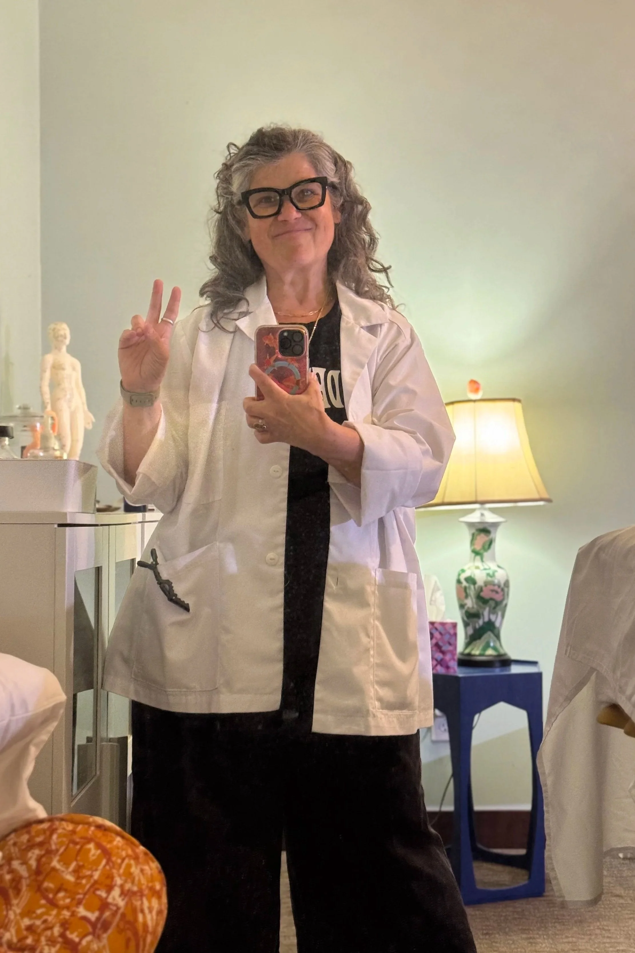 Krista McCain wearing a lab coat at Hundred Grasses Acupuncture, taking a selfie in a treatment room