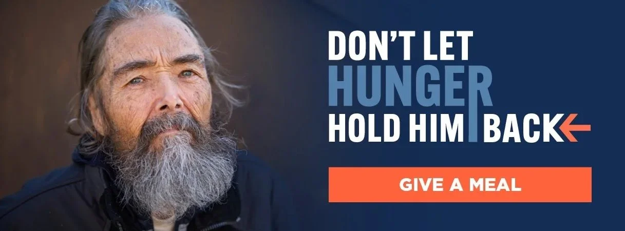 A homeless man with a gray beard and long hair, dressed in a dark jacket, looking directly at the camera. The image is part of an awareness campaign with text that says, 'Don't let hunger hold him back' and a button that says 'Give a meal'.