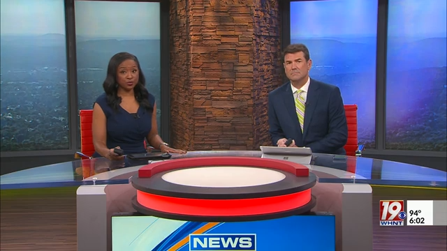 Two news anchors, a woman and a man, sit behind a news desk with a cityscape background and a red and white circular graphic on the desk. The woman is wearing a navy blouse, and the man is in a dark suit with a yellow striped tie. There is a news station logo with the number 19 and temperature and time displayed in the lower right corner.