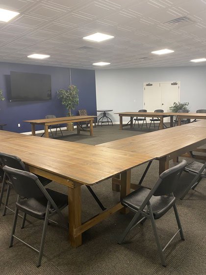 A conference room with a large U-shaped wooden table, black chairs, a wall-mounted TV, a potted plant, and a white door in the background.