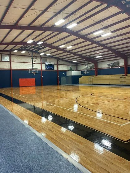Inside an empty gymnasium with a wooden basketball court, blue walls, yellow bleachers, and basketball hoops.