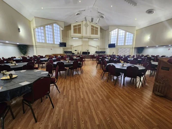 Event hall with round tables covered in black tablecloths, decorated with small centerpieces, and surrounded by red cushioned chairs. The room has wooden floors, high ceilings, large windows, and a stage at the front with audio equipment.