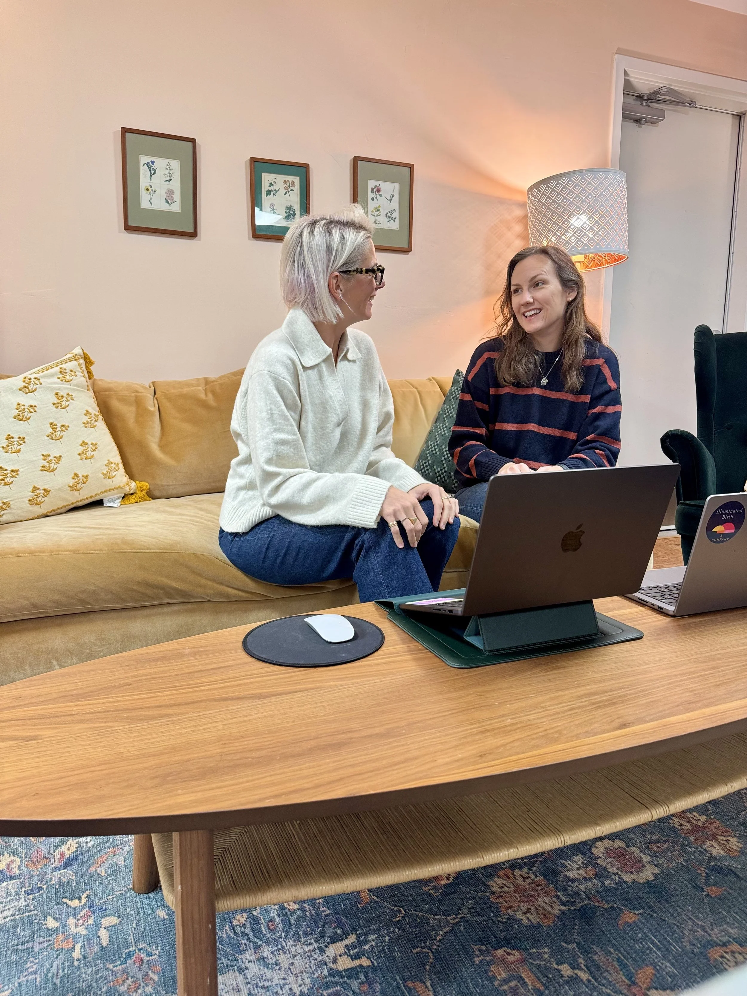Two women sitting on a couch smiling at each other during a Good Company co-working session at Illuminated Birth & Company.