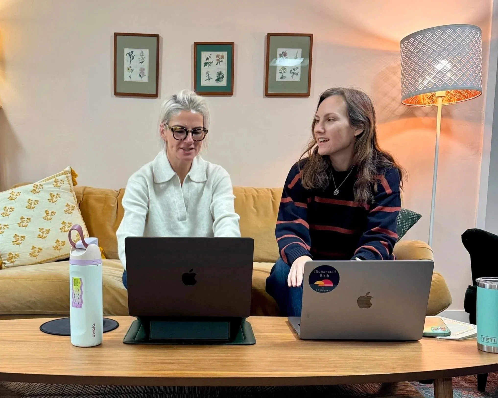 Two women sitting next to each other co-working on their computers on a yellow couch.
