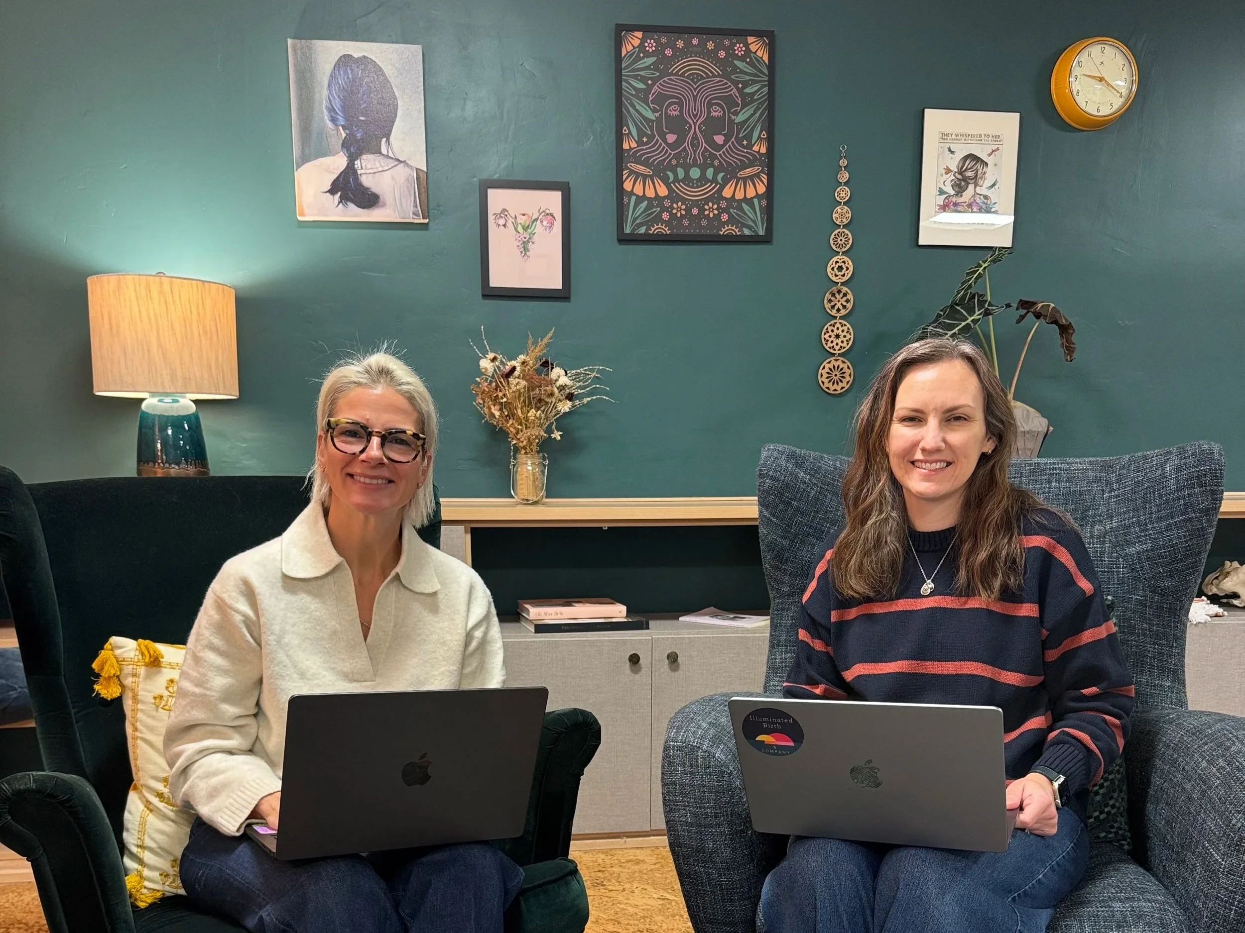Lindsay Binette and Megan Peasley sitting in two arm chairs, smiling at the camera with their laptops in their laps at Illuminated Birth & Company during a co-working session.