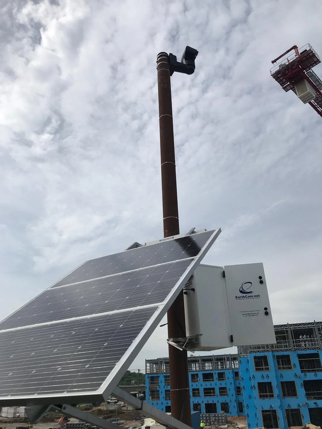 Solar panels mounted on a pole with a chimney and building under construction in the background, under a cloudy sky.