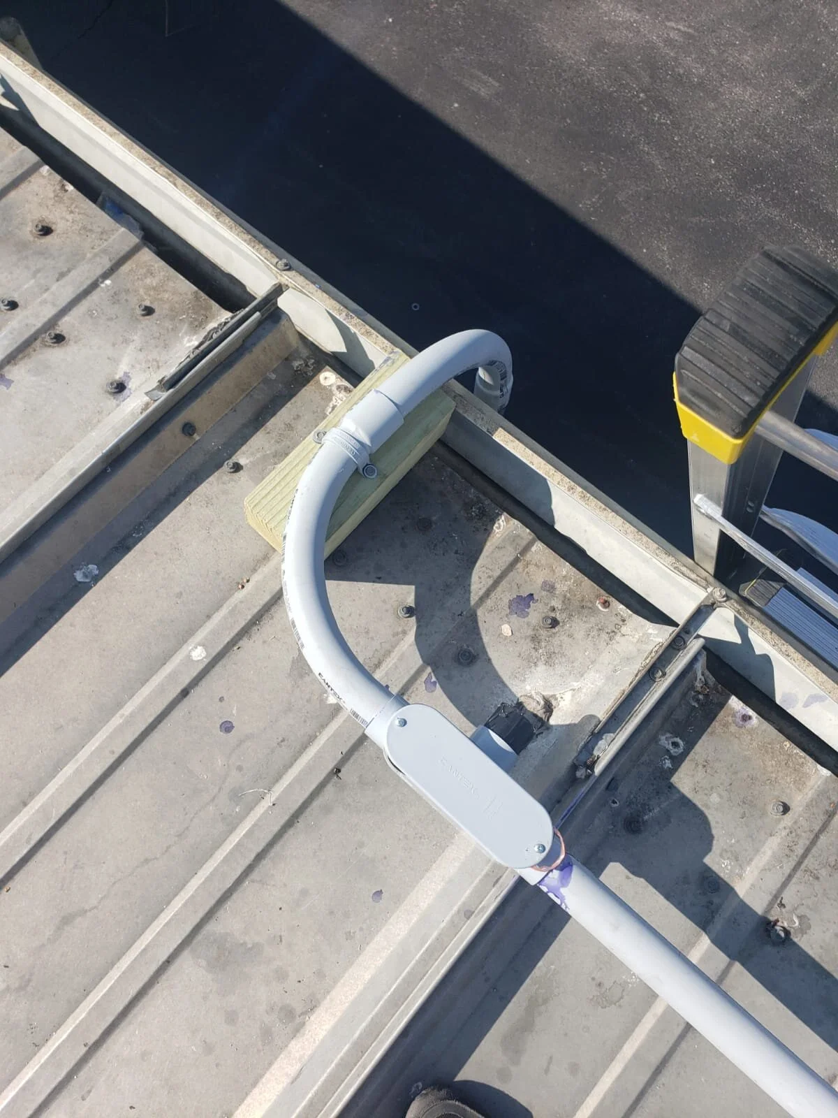 Close-up view of a ladder, pipe, and assembly on a rooftop, with construction materials and an asphalt surface visible.