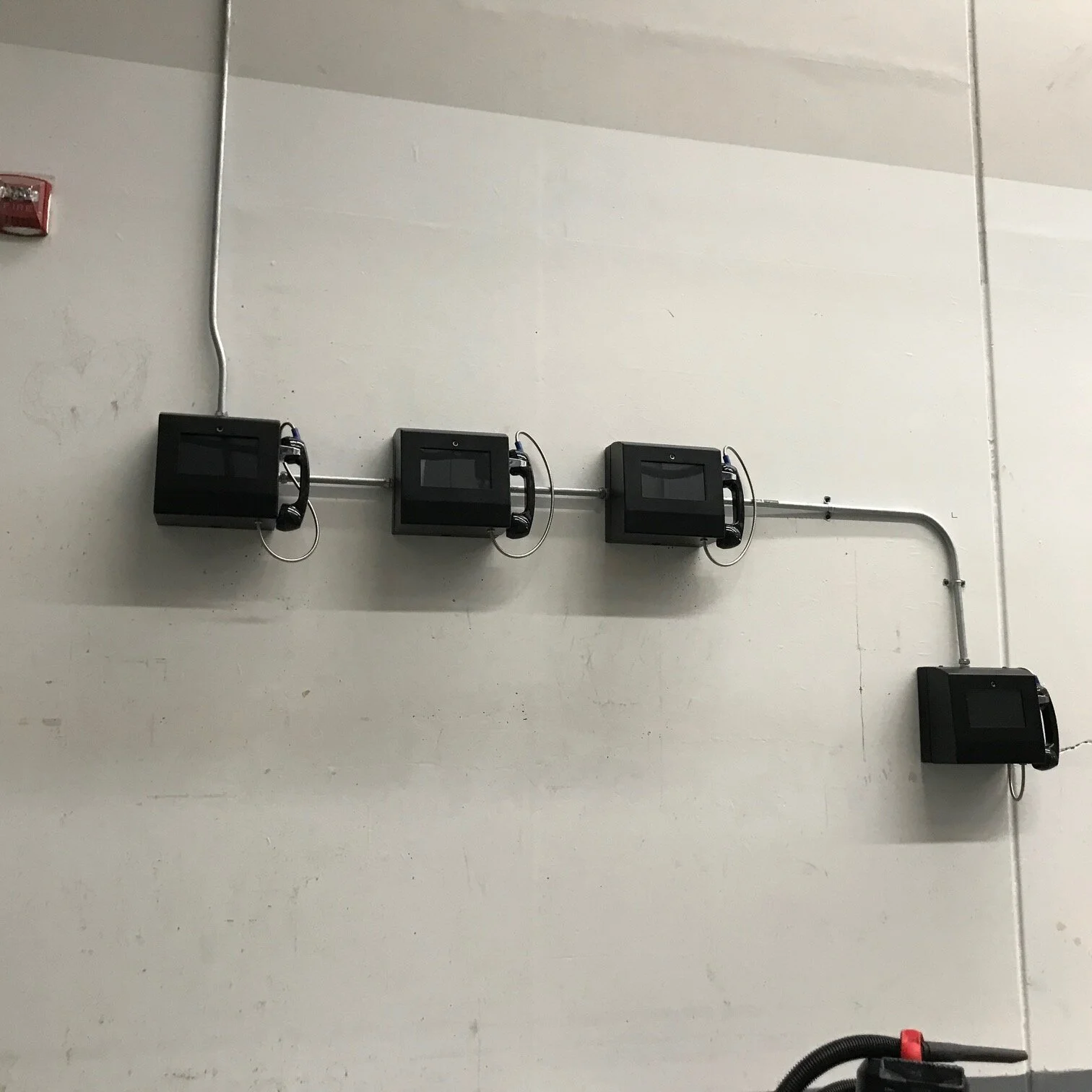 Three black wall-mounted boxes with telephones attached are connected by metal conduits on a white wall. Additionally, there is a small black box with a hose in the bottom right corner.