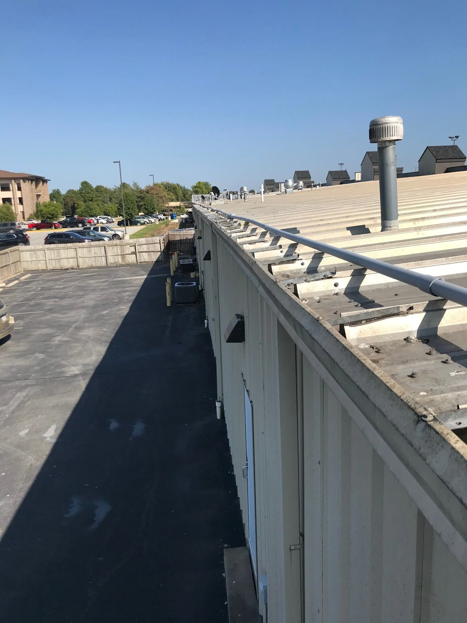 View of a rooftop with ventilators and a metal railing, overlooking a parking lot and green trees in the distance under a clear blue sky.
