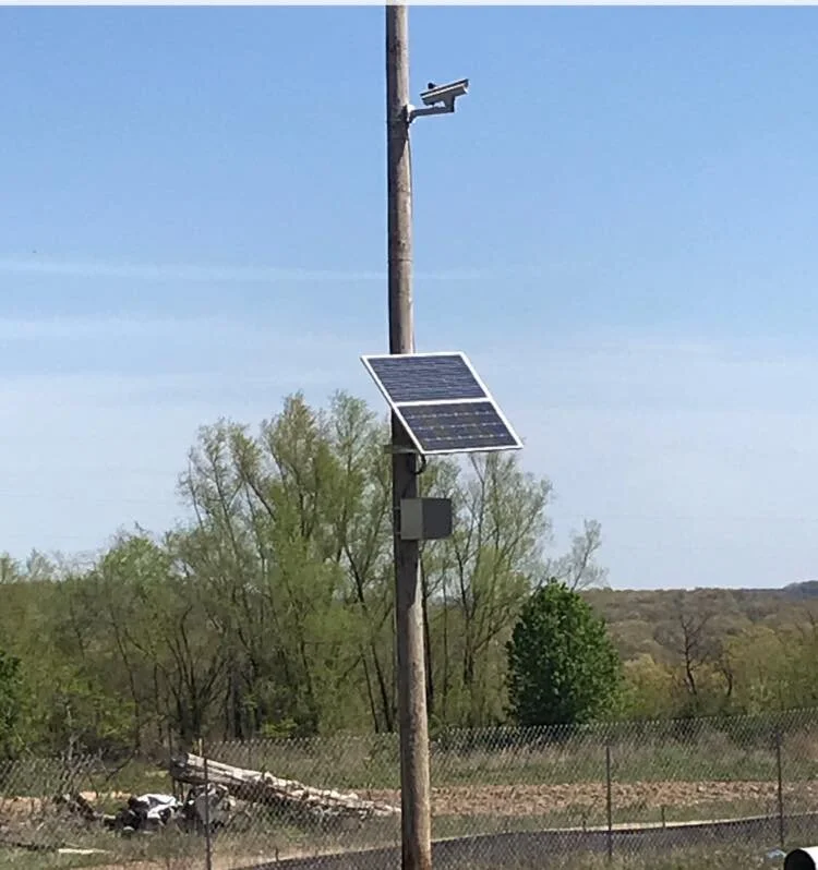 A tall wooden utility pole with solar panels, a security camera, and other equipment mounted on it, in a rural area with trees and a chain-link fence in the background.