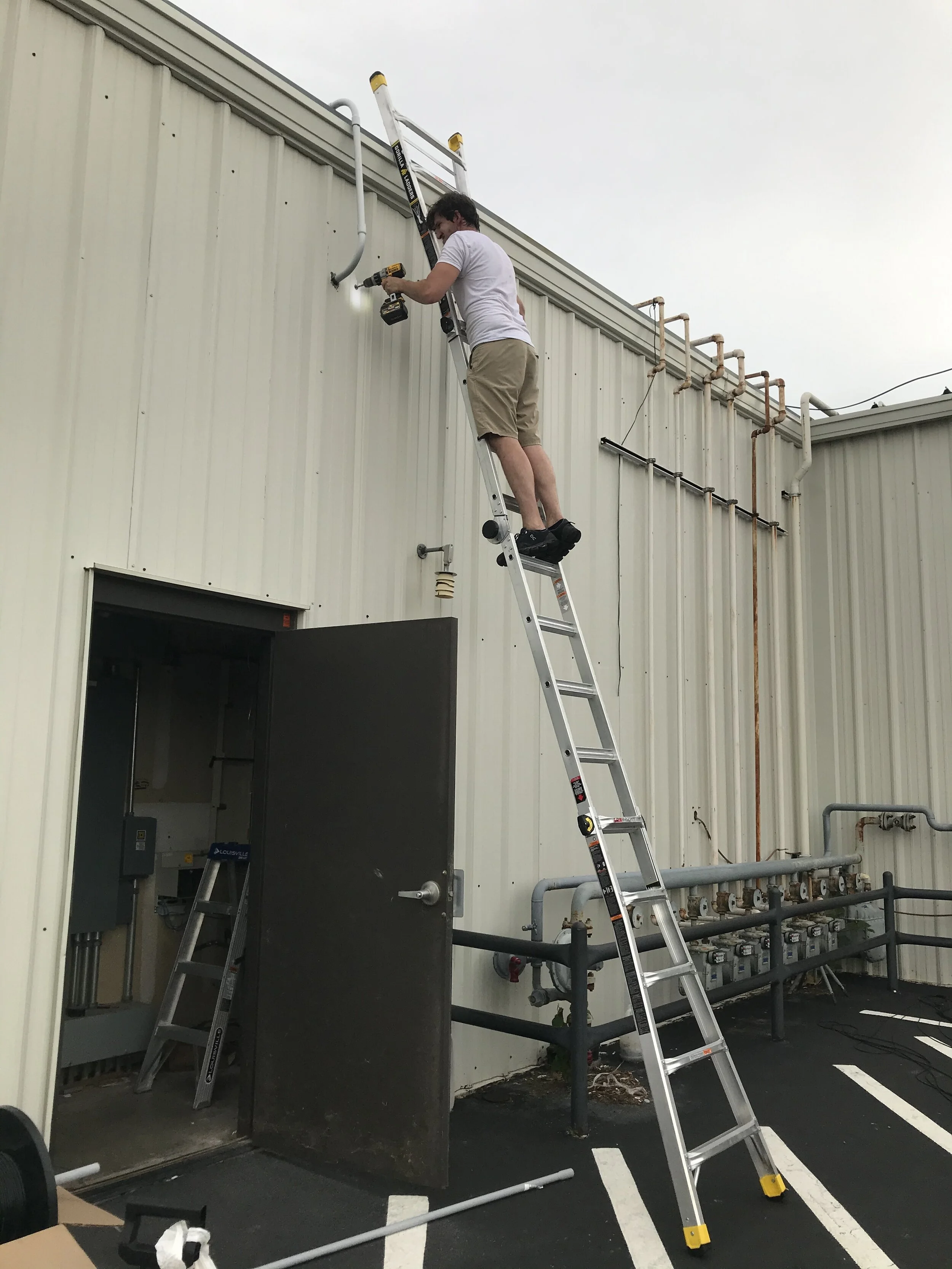 Man on ladder working on pipes outside a building.