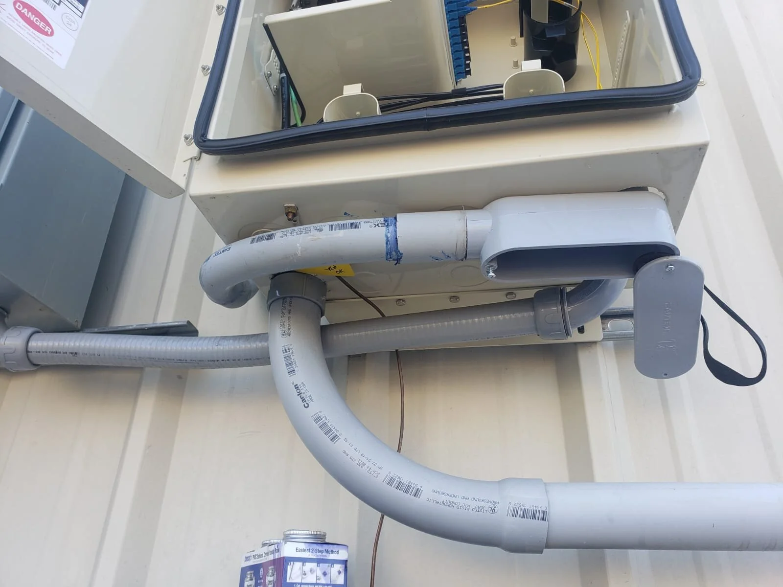 A utility box with electrical components and a transparent cover, showing wiring inside. Several PVC pipes are connected to the box, with one pipe featuring a blue sealant. A warning label and a can of PVC solvent are also visible.