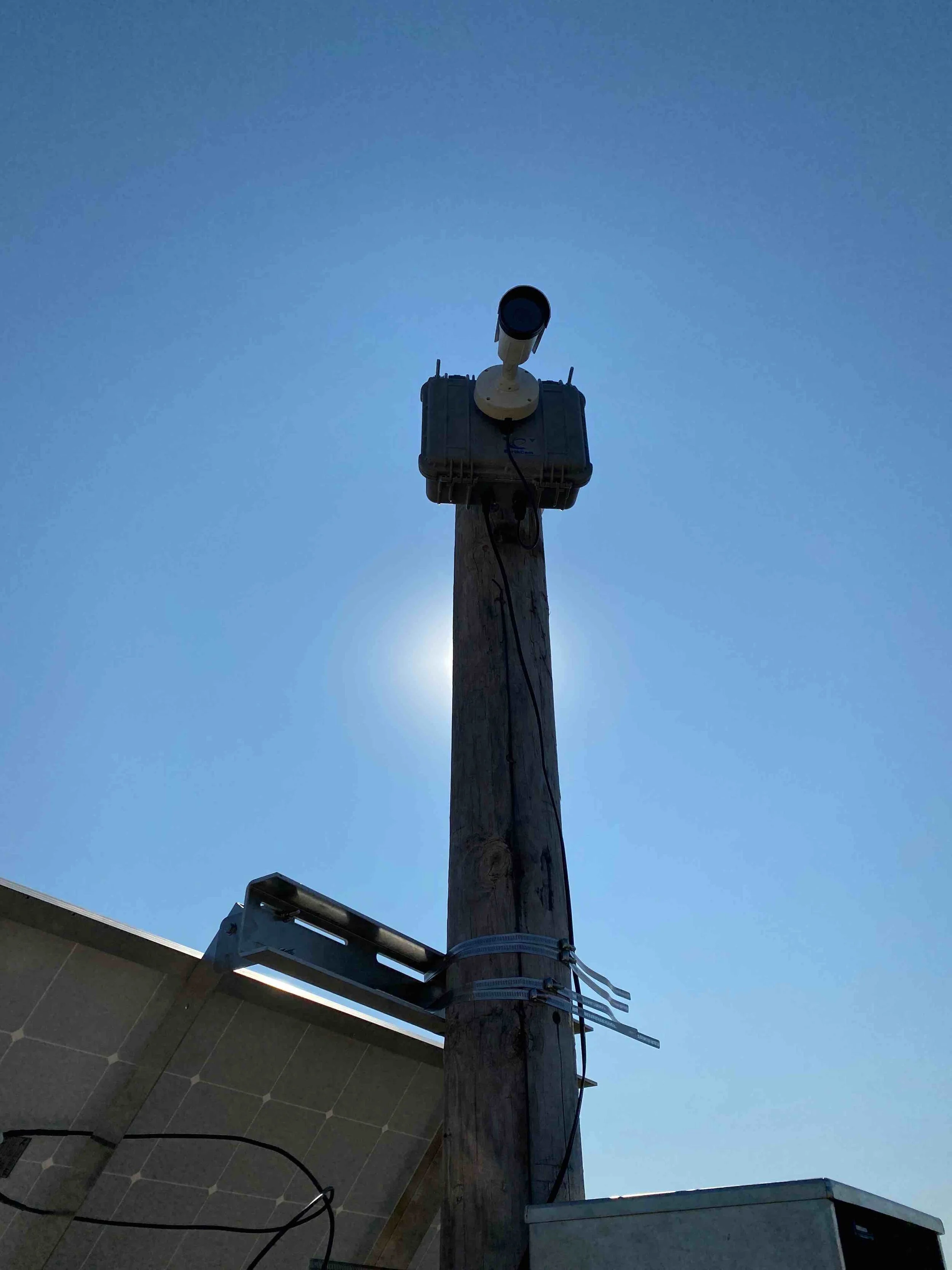 A utility pole with a mounted surveillance camera, electrical box, and solar panel under a clear blue sky.