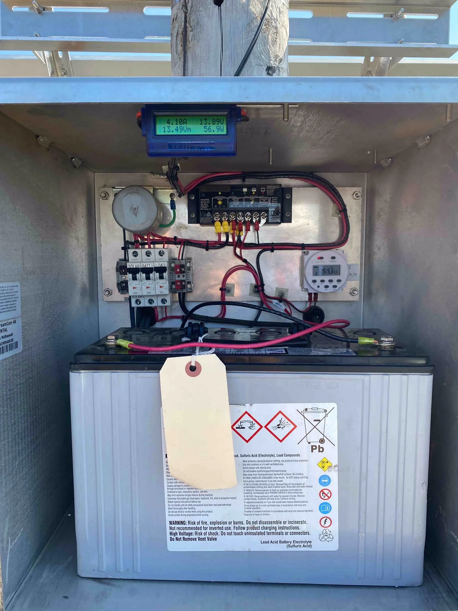 A lead acid battery enclosed in a metal cabinet with wiring, a monitoring device, and warning labels. The battery has a white exterior with hazard symbols and a warning label with instructions and safety precautions.