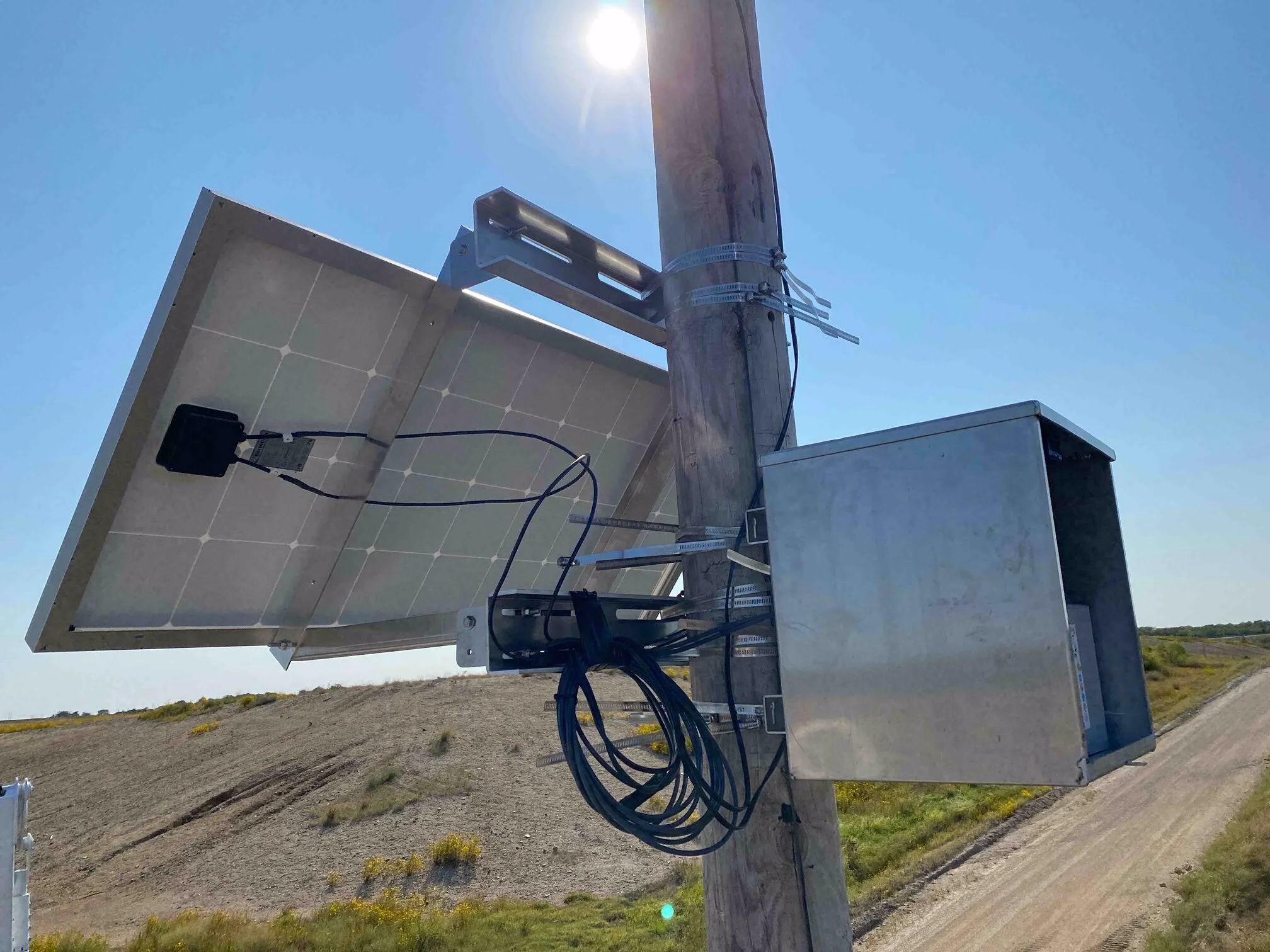 A solar panel mounted on a wooden utility pole with visible wiring and a metal bracket against a clear blue sky, with a bright sun overhead.