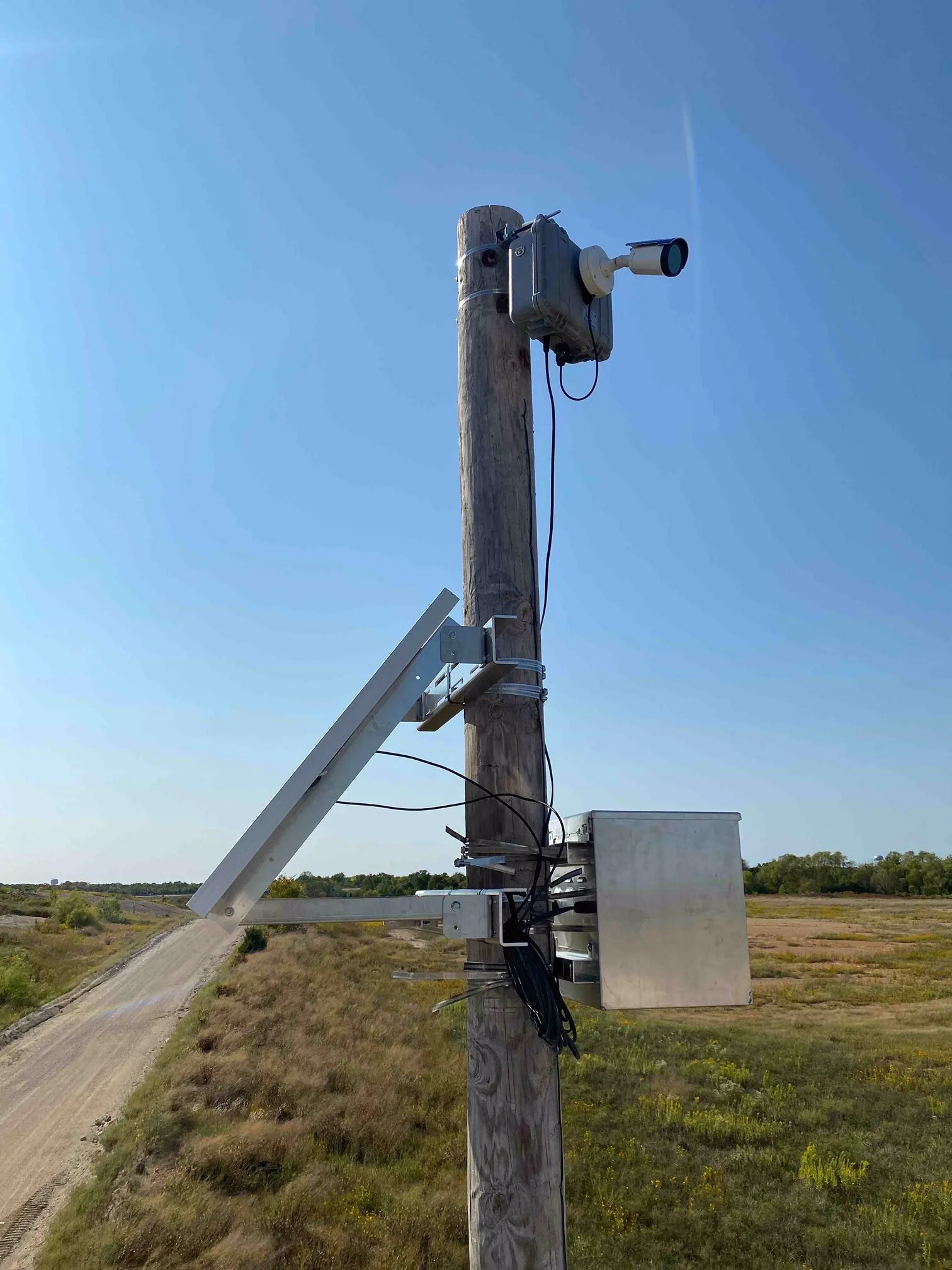 A utility pole with a weather monitoring device and a CCTV camera attached, situated in an open rural area with a dirt road and grassy fields underneath a clear blue sky.