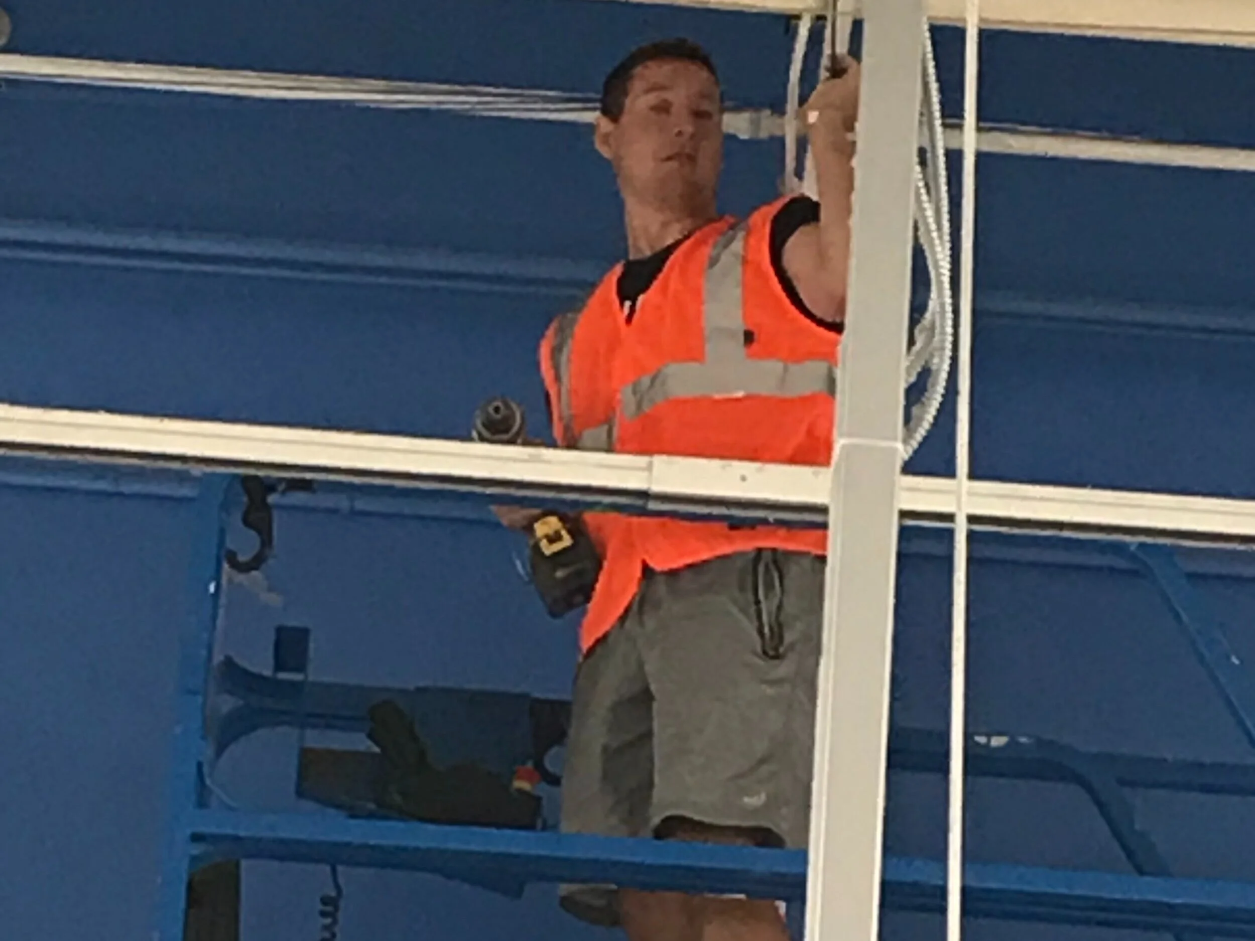 A man is working on maintenance or construction on a tall structure, wearing a bright orange safety vest and a tool belt, against a blue background.