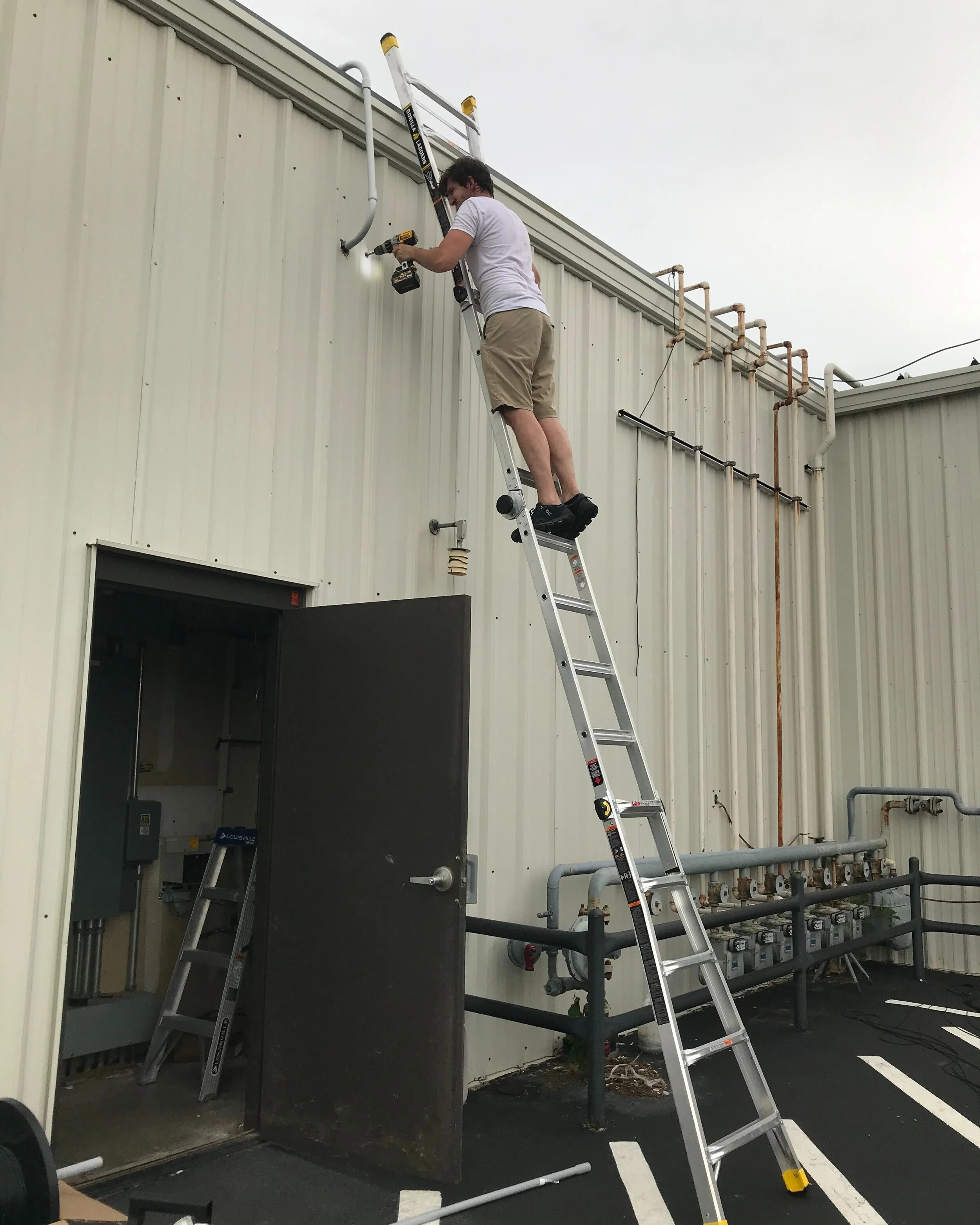 Man standing on ladder working on pipes on a building exterior.