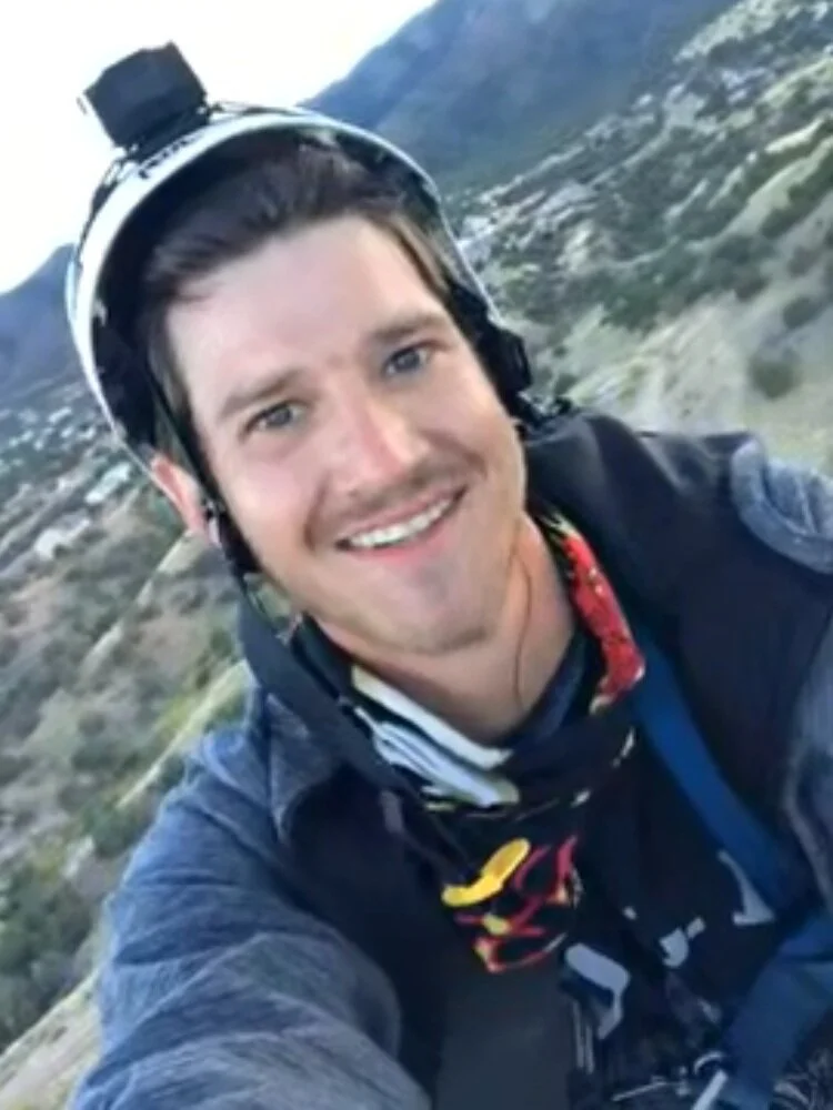 A man smiling while wearing a helmet and outdoor gear, taking a selfie on a hillside with mountains in the background.