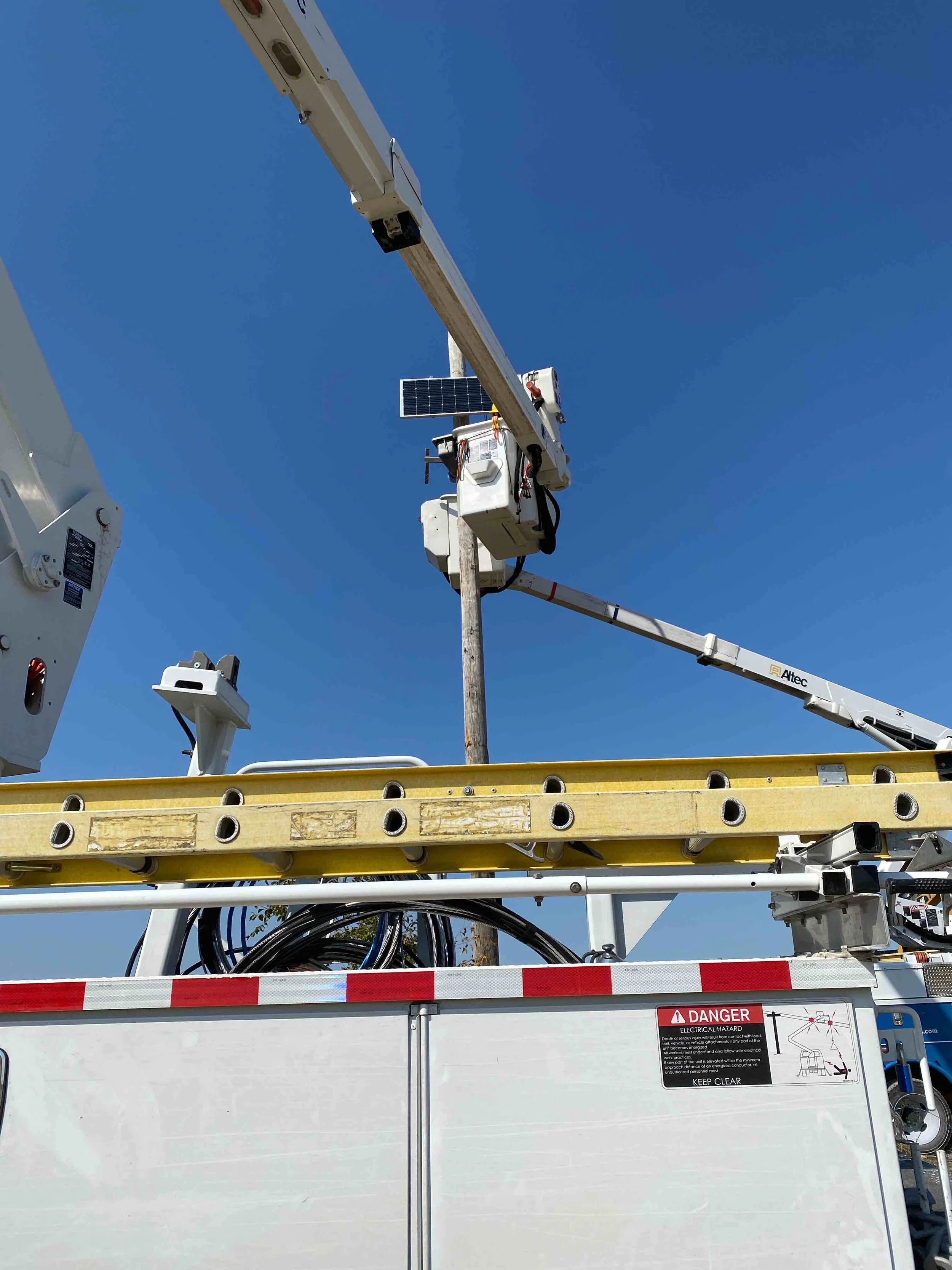 A bucket truck with an extendable arm is parked, with the arm raised and a solar panel attached near the top. The background shows a clear blue sky.