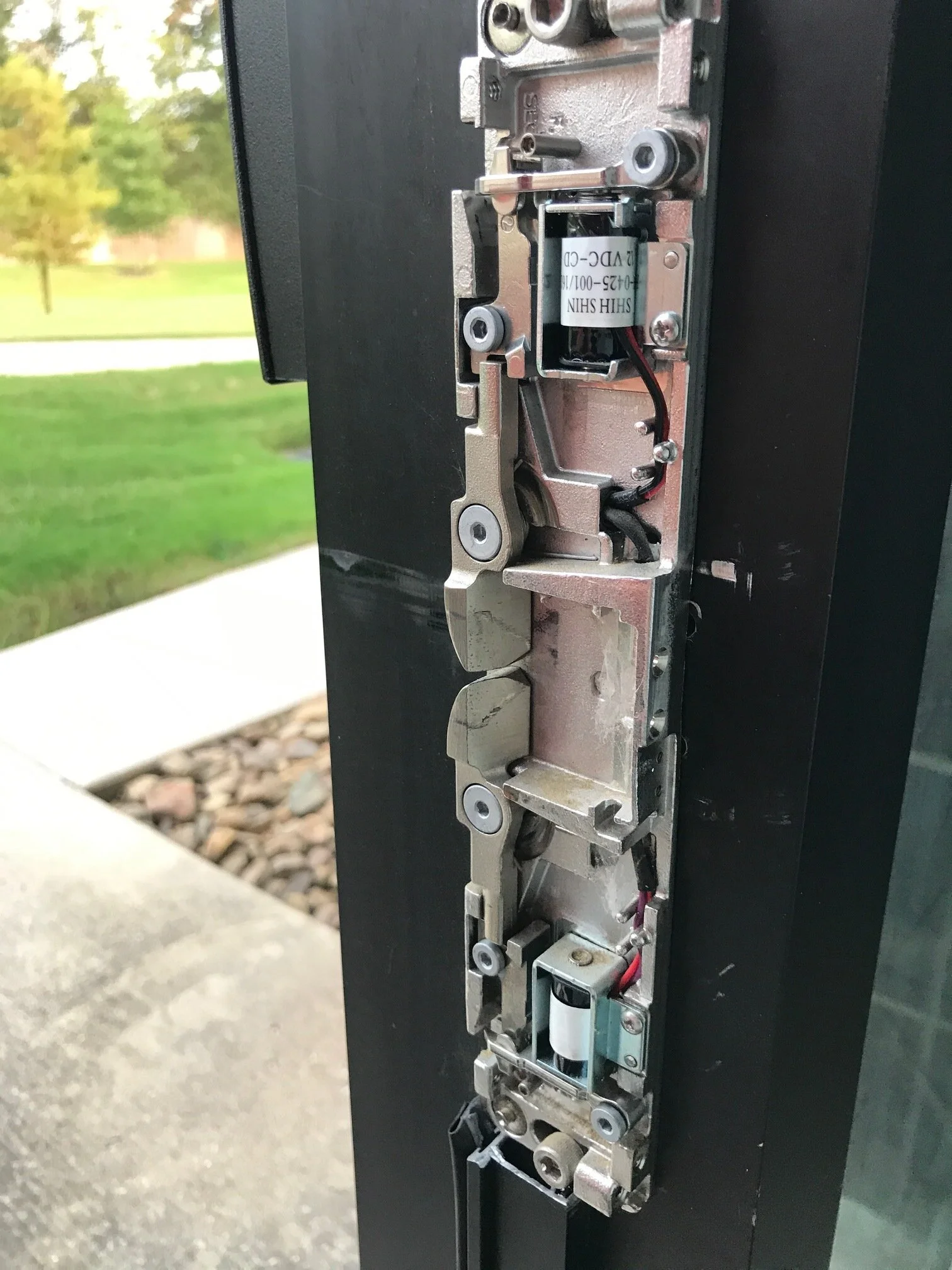 Close-up of a mechanical lock mechanism installed on a black door, with a background of grass and trees outside.