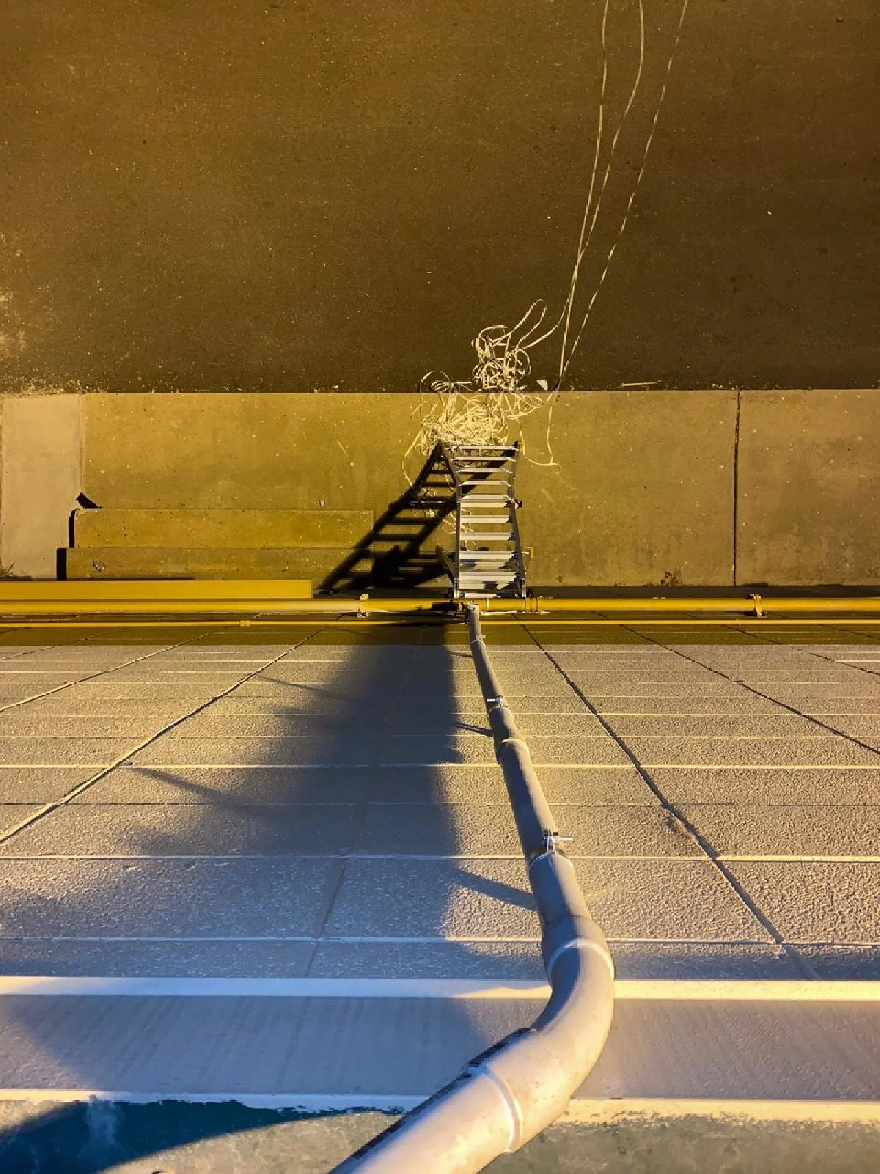 View from a rooftop of a ladder leaning against the edge with a long pipe extending from the ground to the rooftop, cast shadows on the surface below, with a dark sky in the background.